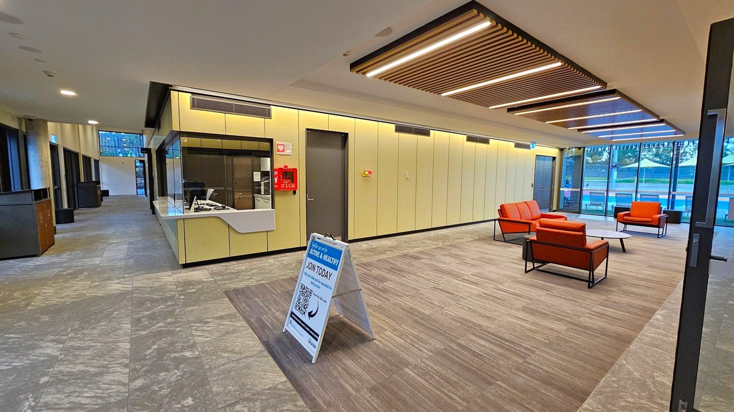A modern interior space with a reception area on the left featuring a counter and a small window. A signboard stands in front of it. The right side has a seating area with orange chairs and a low table on a carpeted section. Large windows in the background reveal an outdoor area with tables and umbrellas. The ceiling has wooden slats with integrated lighting, and the floor is a mix of tiled and carpeted surfaces. A modern interior space with a reception area on the left featuring a counter and a small window. A signboard stands in front of it. The right side has a seating area with orange chairs and a low table on a carpeted section. Large windows in the background reveal an outdoor area with tables and umbrellas. The ceiling has wooden slats with integrated lighting, and the floor is a mix of tiled and carpeted surfaces.