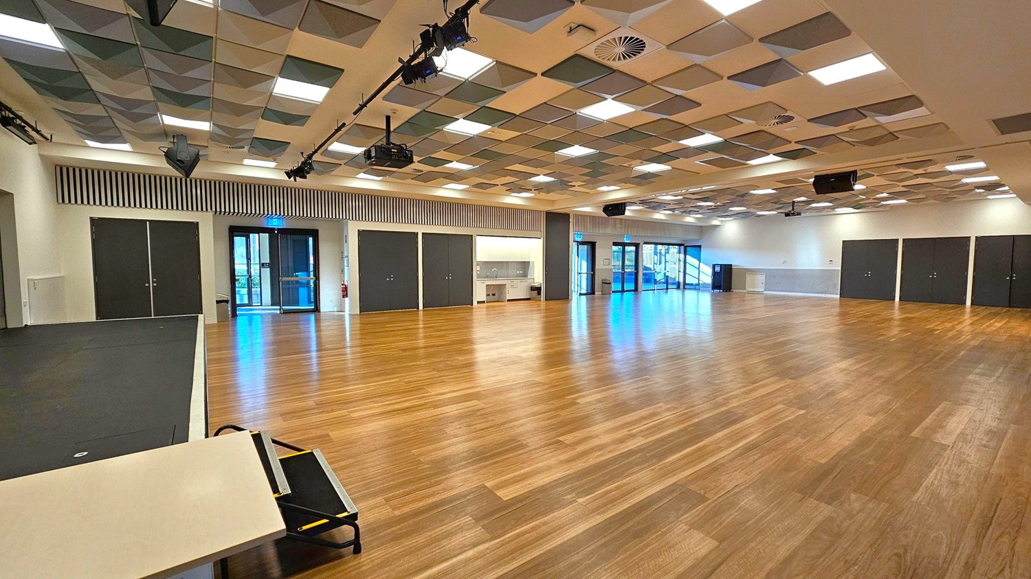 A spacious room with a wooden floor and a patterned ceiling featuring square panels in various shades. On the left, there is a small stage area with a black surface and a table with a folded chair. The back wall has several black double doors and a kitchenette area with a sink. Large glass doors on the right lead outside, allowing natural light to enter. The ceiling has lighting fixtures and speakers, with a projector mounted near the centre. A spacious room with a wooden floor and a patterned ceiling featuring square panels in various shades. On the left, there is a small stage area with a black surface and a table with a folded chair. The back wall has several black double doors and a kitchenette area with a sink. Large glass doors on the right lead outside, allowing natural light to enter. The ceiling has lighting fixtures and speakers, with a projector mounted near the centre.