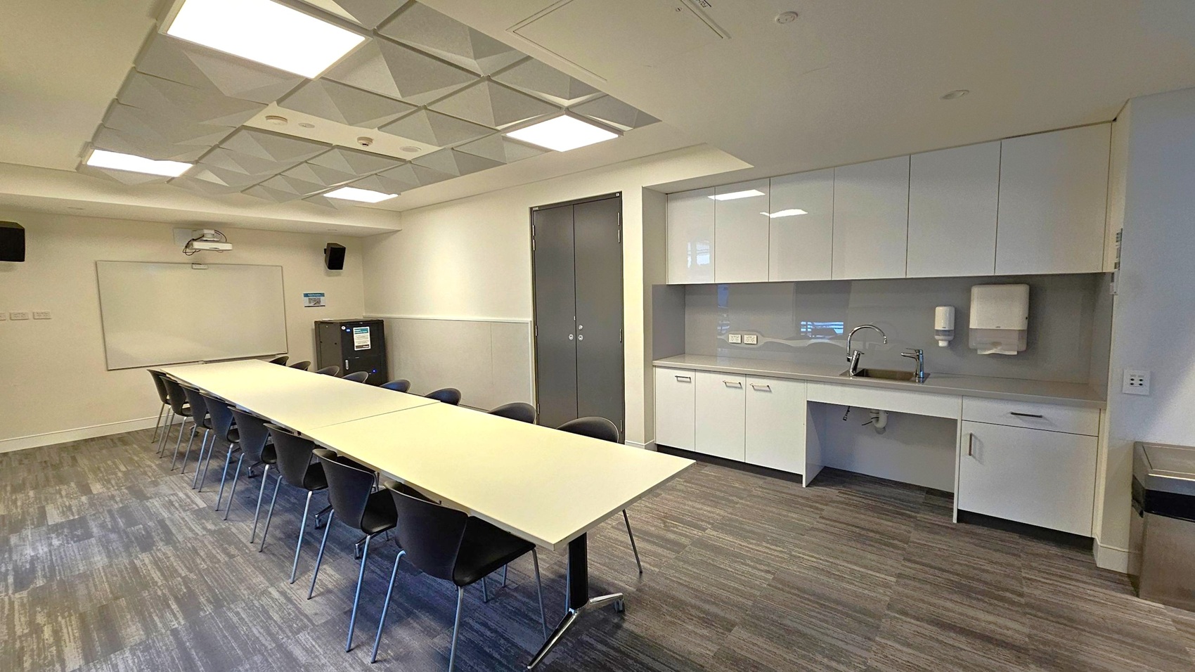 A modern conference room with a long rectangular table in the centre, surrounded by black chairs. On the right, there is a kitchenette with white cabinets, a sink, and a paper towel dispenser. The back wall features a whiteboard and a projector. The ceiling has recessed lighting and a grid pattern. The floor is carpeted with a mix of grey and green tones. A modern conference room with a long rectangular table in the centre, surrounded by black chairs. On the right, there is a kitchenette with white cabinets, a sink, and a paper towel dispenser. The back wall features a whiteboard and a projector. The ceiling has recessed lighting and a grid pattern. The floor is carpeted with a mix of grey and green tones.