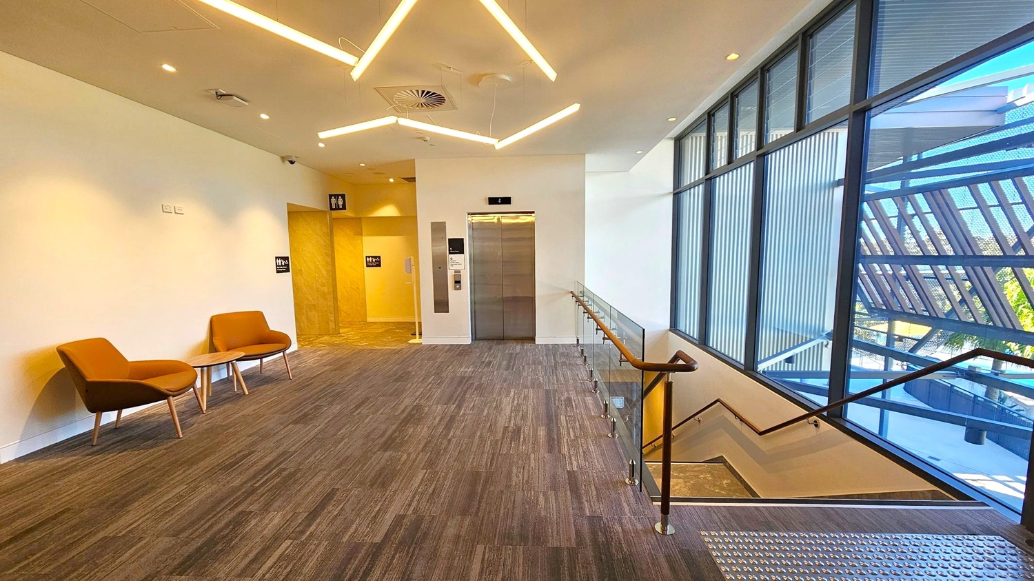 A modern interior space with a grey carpeted floor. On the left, two brown chairs with a small round table are set against a white wall. In the centre, a lift with metallic doors is flanked by signs for restrooms. On the right, a staircase with a wooden handrail descends alongside large windows with a view of an exterior structure. The ceiling features geometric light fixtures and recessed lighting. A modern interior space with a grey carpeted floor. On the left, two brown chairs with a small round table are set against a white wall. In the centre, a lift with metallic doors is flanked by signs for restrooms. On the right, a staircase with a wooden handrail descends alongside large windows with a view of an exterior structure. The ceiling features geometric light fixtures and recessed lighting.