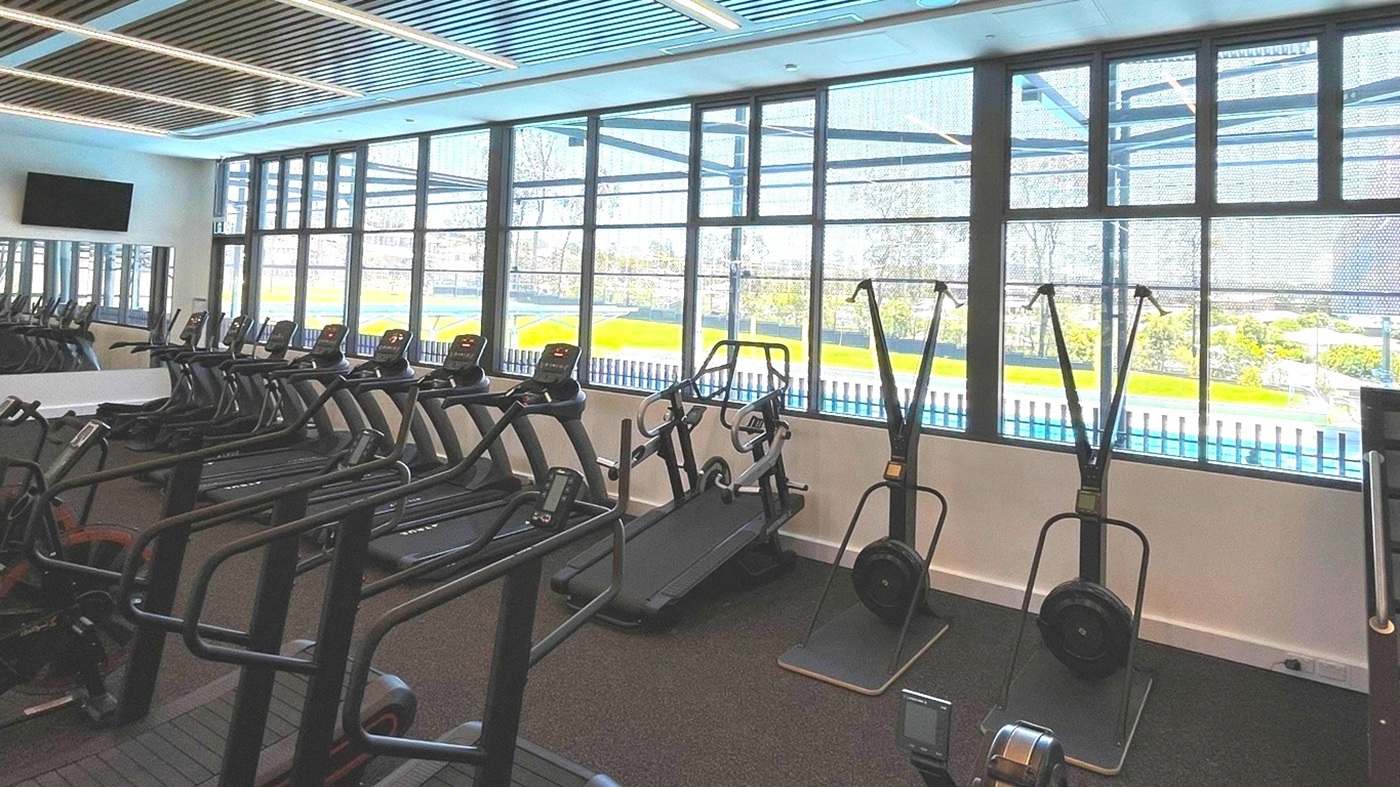 A modern gym interior with a row of treadmills on the left and ski machines in the centre. Large windows on the right provide a view of a green outdoor area and a swimming pool. The ceiling features a grid pattern with integrated lighting. The floor is covered with dark carpeting. A wall-mounted TV is visible on the left. A modern gym interior with a row of treadmills on the left and ski machines in the centre. Large windows on the right provide a view of a green outdoor area and a swimming pool. The ceiling features a grid pattern with integrated lighting. The floor is covered with dark carpeting. A wall-mounted TV is visible on the left.
