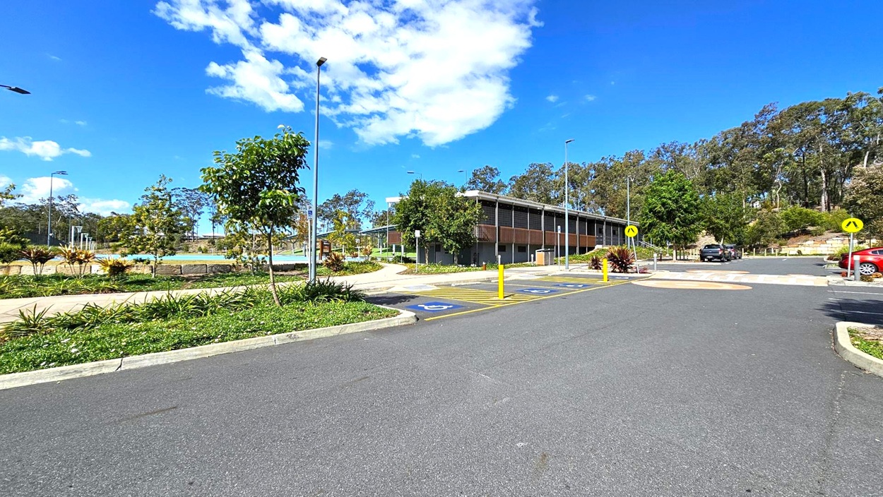 A paved parking area with marked spaces, including disabled parking, is in the foreground. On the left, there are landscaped gardens with small trees and shrubs. In the centre, a modern building with a flat roof and wooden accents is visible. To the right, more parking spaces and a pedestrian crossing are present. The background features a line of tall trees under a clear blue sky with some clouds. A paved parking area with marked spaces, including disabled parking, is in the foreground. On the left, there are landscaped gardens with small trees and shrubs. In the centre, a modern building with a flat roof and wooden accents is visible. To the right, more parking spaces and a pedestrian crossing are present. The background features a line of tall trees under a clear blue sky with some clouds.