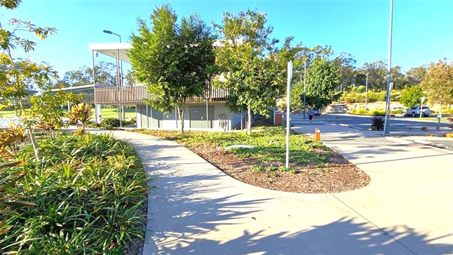 A paved pathway curves through a landscaped area with green shrubs and small trees on both sides. In the background, a modern building with a flat roof and vertical slats is partially visible. The area is well-maintained, with a streetlamp and a few cars parked in the distance. The sky is clear, and the shadows suggest it is a sunny day. A paved pathway curves through a landscaped area with green shrubs and small trees on both sides. In the background, a modern building with a flat roof and vertical slats is partially visible. The area is well-maintained, with a streetlamp and a few cars parked in the distance. The sky is clear, and the shadows suggest it is a sunny day.