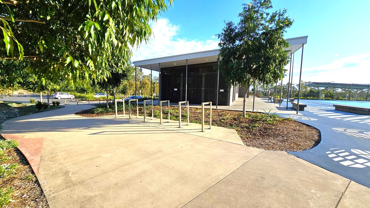 A modern building with a flat roof is situated in the centre, surrounded by a paved area. On the left, there are metal bike racks and a path leading to a car park with a few vehicles visible. Trees and shrubs are planted around the area, providing greenery. On the right, a blue pathway with white patterns runs alongside the building, leading to a covered area with benches. The sky is clear with some clouds. A modern building with a flat roof is situated in the centre, surrounded by a paved area. On the left, there are metal bike racks and a path leading to a car park with a few vehicles visible. Trees and shrubs are planted around the area, providing greenery. On the right, a blue pathway with white patterns runs alongside the building, leading to a covered area with benches. The sky is clear with some clouds.