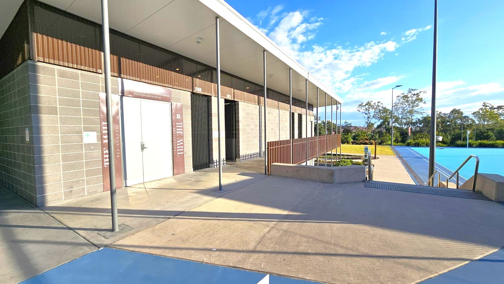 A modern building with a flat roof and a facade of grey bricks and wooden panels on the left. A large door with signage is visible. Several metal poles support the overhanging roof. On the right, there is a swimming pool with clear blue water, bordered by a concrete walkway. In the background, there are trees and a grassy area. The ground is paved with concrete, and the ceiling is flat and white. A modern building with a flat roof and a facade of grey bricks and wooden panels on the left. A large door with signage is visible. Several metal poles support the overhanging roof. On the right, there is a swimming pool with clear blue water, bordered by a concrete walkway. In the background, there are trees and a grassy area. The ground is paved with concrete, and the ceiling is flat and white.