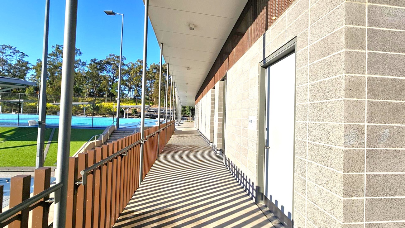 A walkway with a wooden fence on the left and a brick wall on the right. The floor is made of wooden planks, and the ceiling is flat and white. There are tall metal poles supporting the structure. Beyond the fence, there is a blue sports court and green grass. Trees are visible in the background under a clear blue sky. A walkway with a wooden fence on the left and a brick wall on the right. The floor is made of wooden planks, and the ceiling is flat and white. There are tall metal poles supporting the structure. Beyond the fence, there is a blue sports court and green grass. Trees are visible in the background under a clear blue sky.