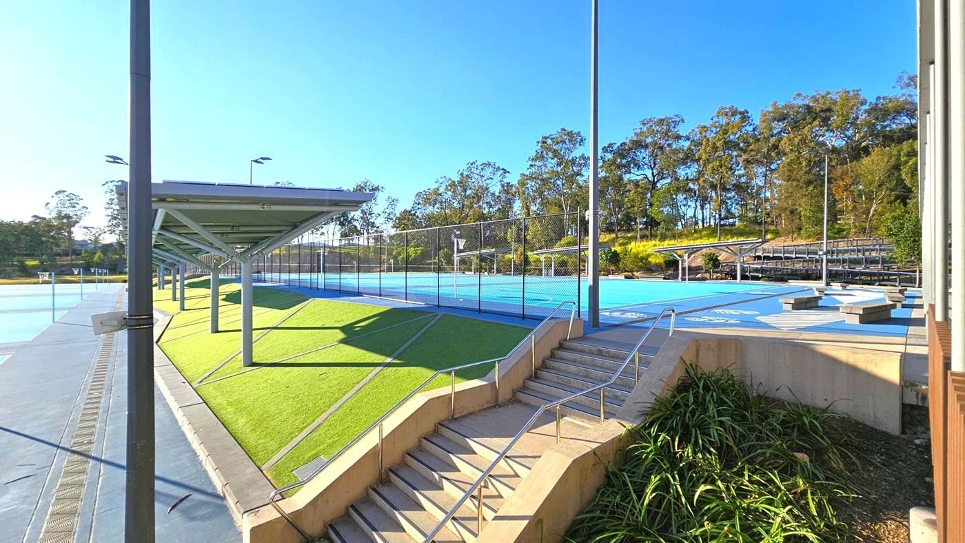 On the left, there is a covered walkway with a metal roof supported by poles, adjacent to a green artificial turf area. In the centre, a blue sports court is enclosed by a chain-link fence. On the right, a set of concrete steps with metal railings leads up to the court, with some plants at the base. In the background, there are trees and a small set of bleachers. The sky is clear and blue. On the left, there is a covered walkway with a metal roof supported by poles, adjacent to a green artificial turf area. In the centre, a blue sports court is enclosed by a chain-link fence. On the right, a set of concrete steps with metal railings leads up to the court, with some plants at the base. In the background, there are trees and a small set of bleachers. The sky is clear and blue.