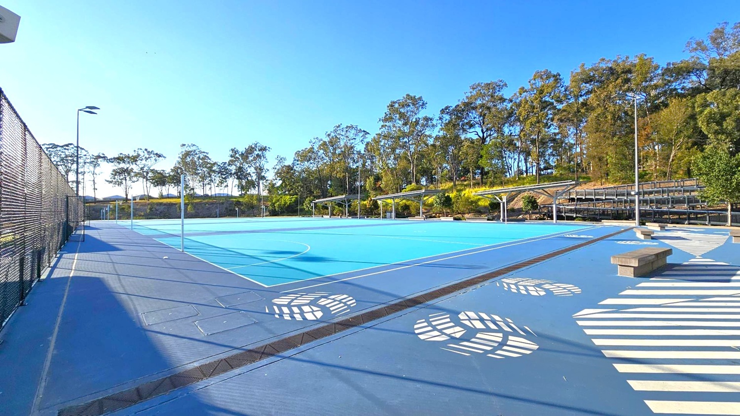 An outdoor sports court with a blue surface and white markings. On the left, a tall black chain-link fence runs alongside the court. In the background, there are trees and a small hill. On the right, there are covered seating areas with metal benches. The foreground features a patterned walkway with white designs and a few concrete benches. The area is well-lit with tall floodlights. An outdoor sports court with a blue surface and white markings. On the left, a tall black chain-link fence runs alongside the court. In the background, there are trees and a small hill. On the right, there are covered seating areas with metal benches. The foreground features a patterned walkway with white designs and a few concrete benches. The area is well-lit with tall floodlights.