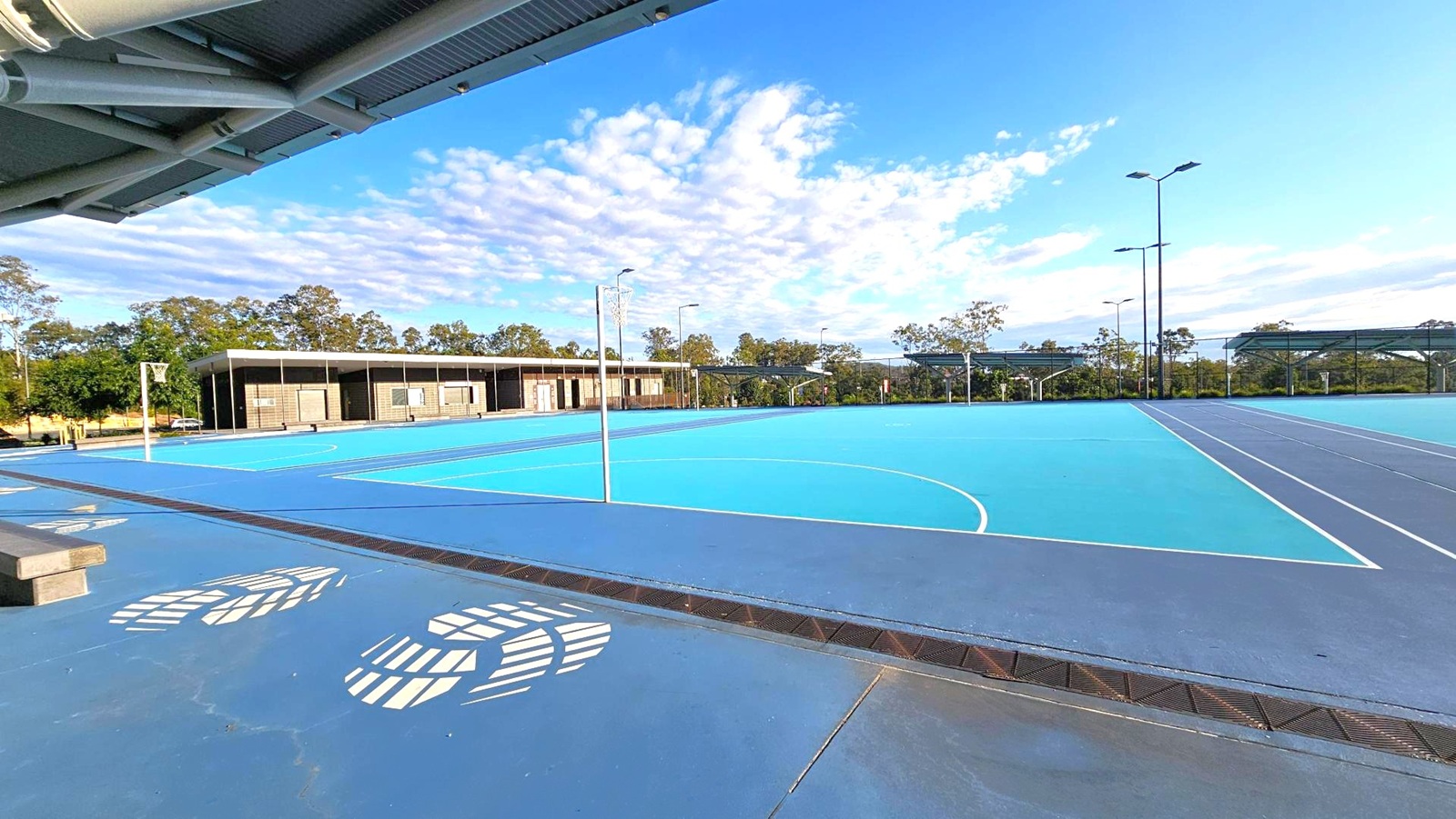 Outdoor sports courts with blue surfaces and white lines, featuring netball hoops. On the left, a covered seating area with a metal roof and large white footprints painted on the ground. In the background, a row of modern, dark-coloured buildings with large windows. Trees and greenery are visible beyond the buildings, and tall floodlights are positioned around the courts. Outdoor sports courts with blue surfaces and white lines, featuring netball hoops. On the left, a covered seating area with a metal roof and large white footprints painted on the ground. In the background, a row of modern, dark-coloured buildings with large windows. Trees and greenery are visible beyond the buildings, and tall floodlights are positioned around the courts.