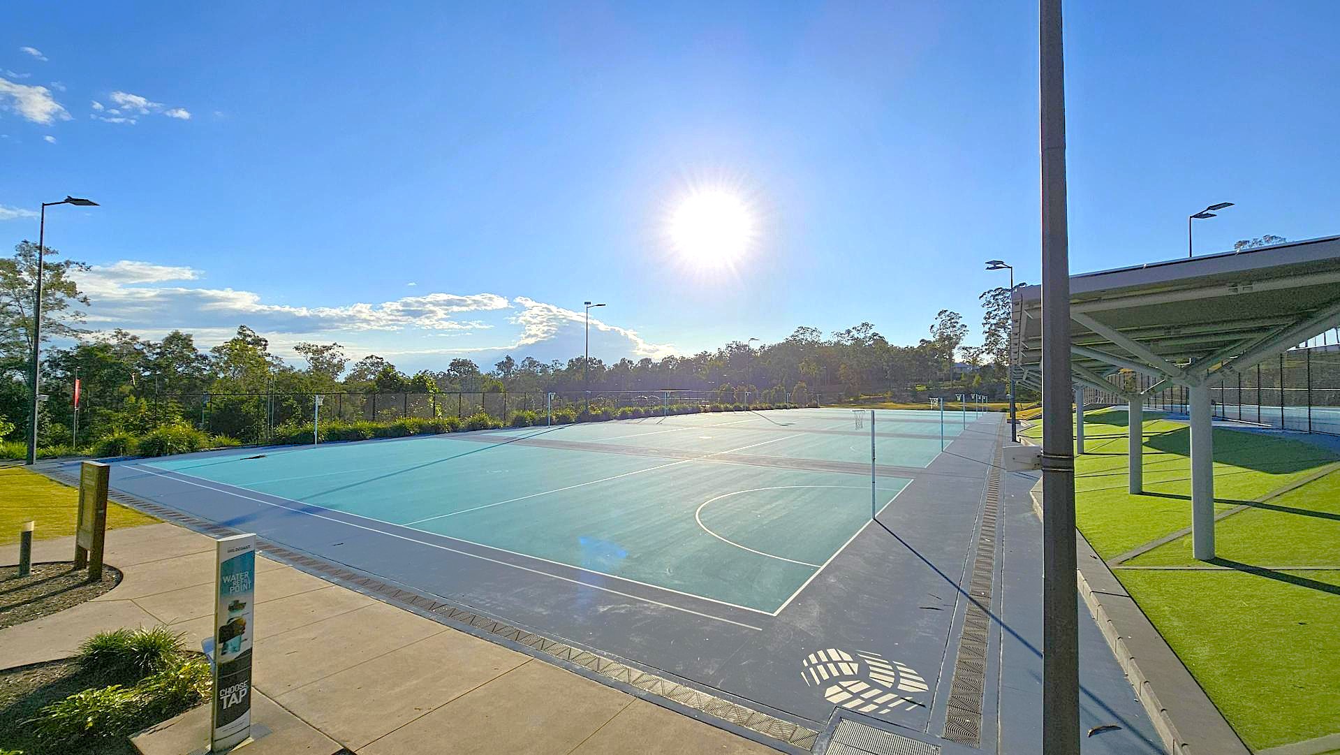 A sports court with a blue surface is in the centre, surrounded by a fence. On the left, there's a concrete path with a water station and some greenery. To the right, a covered area with a green artificial turf is supported by metal poles. The background features trees and a clear sky with the sun shining brightly. A sports court with a blue surface is in the centre, surrounded by a fence. On the left, there's a concrete path with a water station and some greenery. To the right, a covered area with a green artificial turf is supported by metal poles. The background features trees and a clear sky with the sun shining brightly.