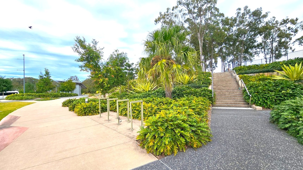A landscaped outdoor area with a pathway on the left leading to a bike rack. Lush green bushes and small trees line the path. On the right, a set of stairs with metal railings ascends through dense foliage. Tall trees are visible in the background. The ground is a mix of smooth concrete and textured surfaces. A landscaped outdoor area with a pathway on the left leading to a bike rack. Lush green bushes and small trees line the path. On the right, a set of stairs with metal railings ascends through dense foliage. Tall trees are visible in the background. The ground is a mix of smooth concrete and textured surfaces.