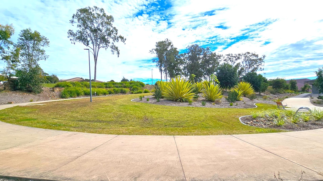 A landscaped park area with a curved concrete path in the foreground. The middle features a circular garden bed with spiky green plants and small shrubs, surrounded by grass. To the right, a bench is placed near the path. Tall trees are scattered throughout the background, with a few streetlights visible. The sky is partly cloudy, casting a soft light over the scene. A landscaped park area with a curved concrete path in the foreground. The middle features a circular garden bed with spiky green plants and small shrubs, surrounded by grass. To the right, a bench is placed near the path. Tall trees are scattered throughout the background, with a few streetlights visible. The sky is partly cloudy, casting a soft light over the scene.