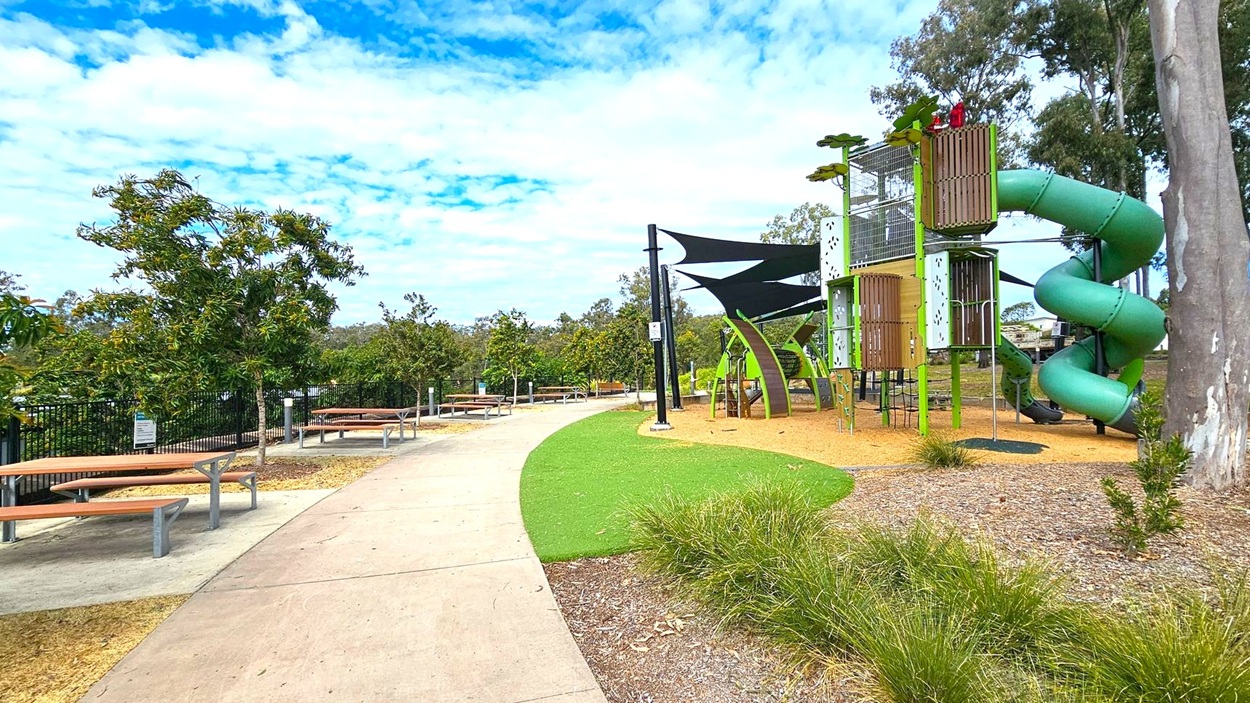 A park scene with a paved path running through the centre. On the left, there are several picnic tables with benches under small trees. On the right, a playground features a tall structure with a green spiral slide and climbing elements. The ground is covered with mulch and artificial grass. In the background, there are more trees and a fence. The sky is partly cloudy. A park scene with a paved path running through the centre. On the left, there are several picnic tables with benches under small trees. On the right, a playground features a tall structure with a green spiral slide and climbing elements. The ground is covered with mulch and artificial grass. In the background, there are more trees and a fence. The sky is partly cloudy.