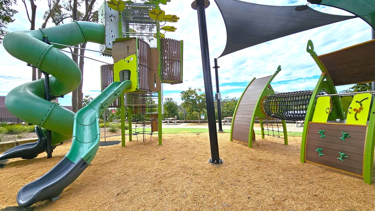 A playground with a large green spiral slide on the left, connected to a tall climbing structure. In the centre, there's a vertical climbing net and a small slide. On the right, a climbing wall and a tunnel made of netting are visible. The ground is covered with wood chips, and there are shade sails overhead. In the background, there are trees and picnic tables on a grassy area. A playground with a large green spiral slide on the left, connected to a tall climbing structure. In the centre, there's a vertical climbing net and a small slide. On the right, a climbing wall and a tunnel made of netting are visible. The ground is covered with wood chips, and there are shade sails overhead. In the background, there are trees and picnic tables on a grassy area.