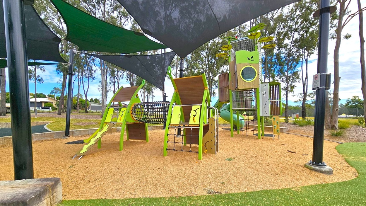 A playground with various climbing structures and slides, primarily in green and brown. The ground is covered with tan mulch, and there are black shade sails overhead. In the background, there are tall trees and a grassy area. A black pole with a sign is visible on the right. The playground equipment includes a net tunnel and a tall tower with a slide. A playground with various climbing structures and slides, primarily in green and brown. The ground is covered with tan mulch, and there are black shade sails overhead. In the background, there are tall trees and a grassy area. A black pole with a sign is visible on the right. The playground equipment includes a net tunnel and a tall tower with a slide.