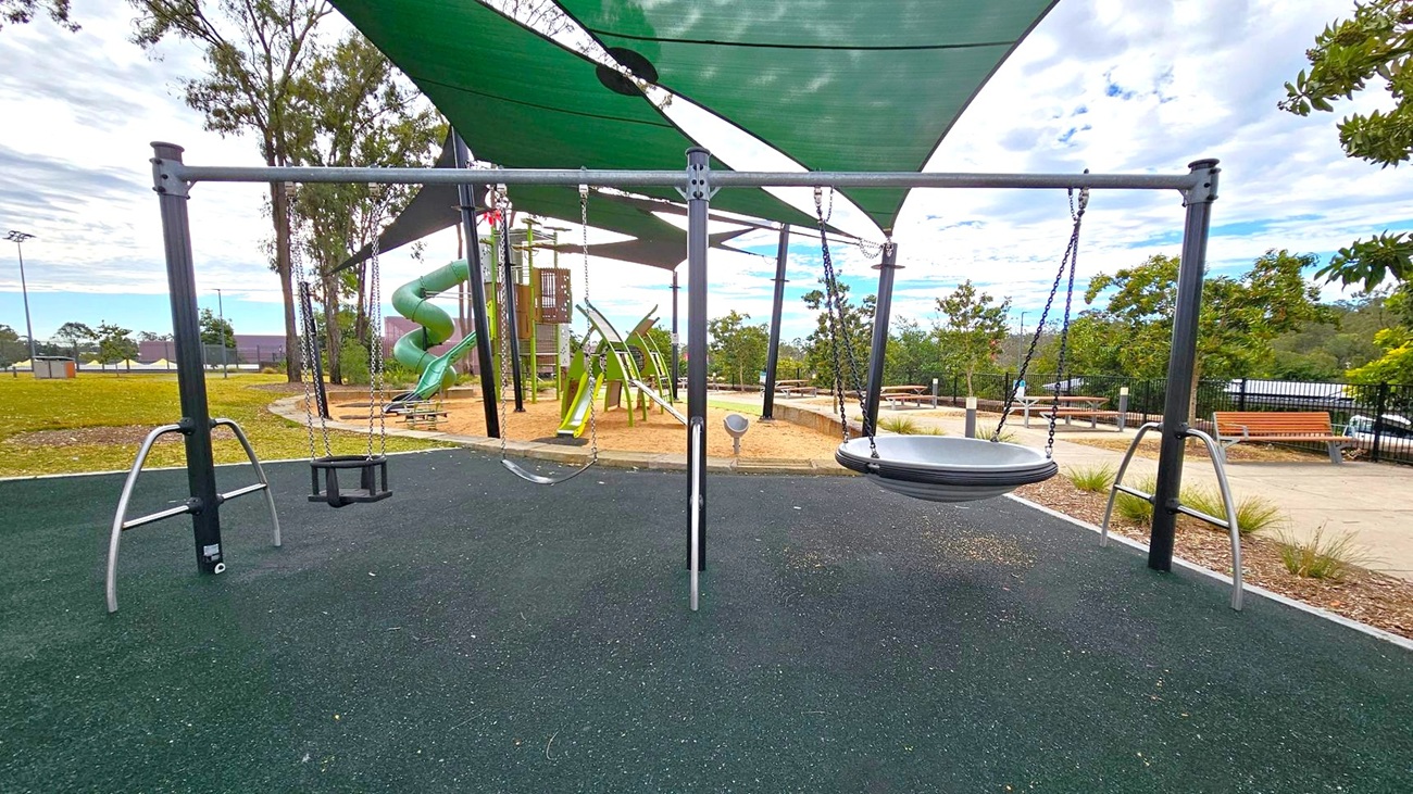 A playground with a swing set in the foreground, featuring a toddler swing and a circular disc swing. The ground is covered with dark rubber matting. In the background, there is a green spiral slide and climbing structures. Green shade sails are overhead, providing cover. To the right, there are benches and a path lined with small plants. Trees and a grassy area are visible in the distance. A playground with a swing set in the foreground, featuring a toddler swing and a circular disc swing. The ground is covered with dark rubber matting. In the background, there is a green spiral slide and climbing structures. Green shade sails are overhead, providing cover. To the right, there are benches and a path lined with small plants. Trees and a grassy area are visible in the distance.