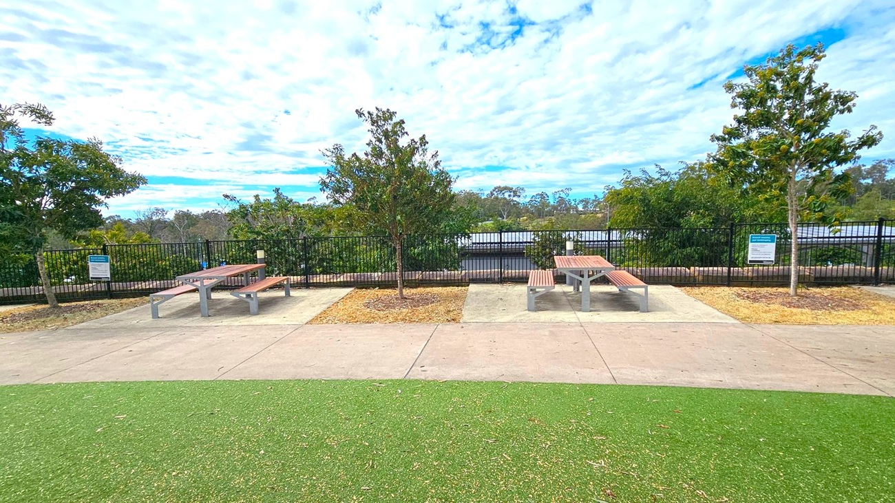 In the foreground, there is a green artificial turf area leading to a concrete path. In the middle, two picnic tables with wooden benches are placed on concrete slabs, separated by small patches of grass with young trees. The background features a black metal fence with dense greenery and trees beyond it. Signs are visible near the tables, and the sky is filled with scattered clouds. In the foreground, there is a green artificial turf area leading to a concrete path. In the middle, two picnic tables with wooden benches are placed on concrete slabs, separated by small patches of grass with young trees. The background features a black metal fence with dense greenery and trees beyond it. Signs are visible near the tables, and the sky is filled with scattered clouds.