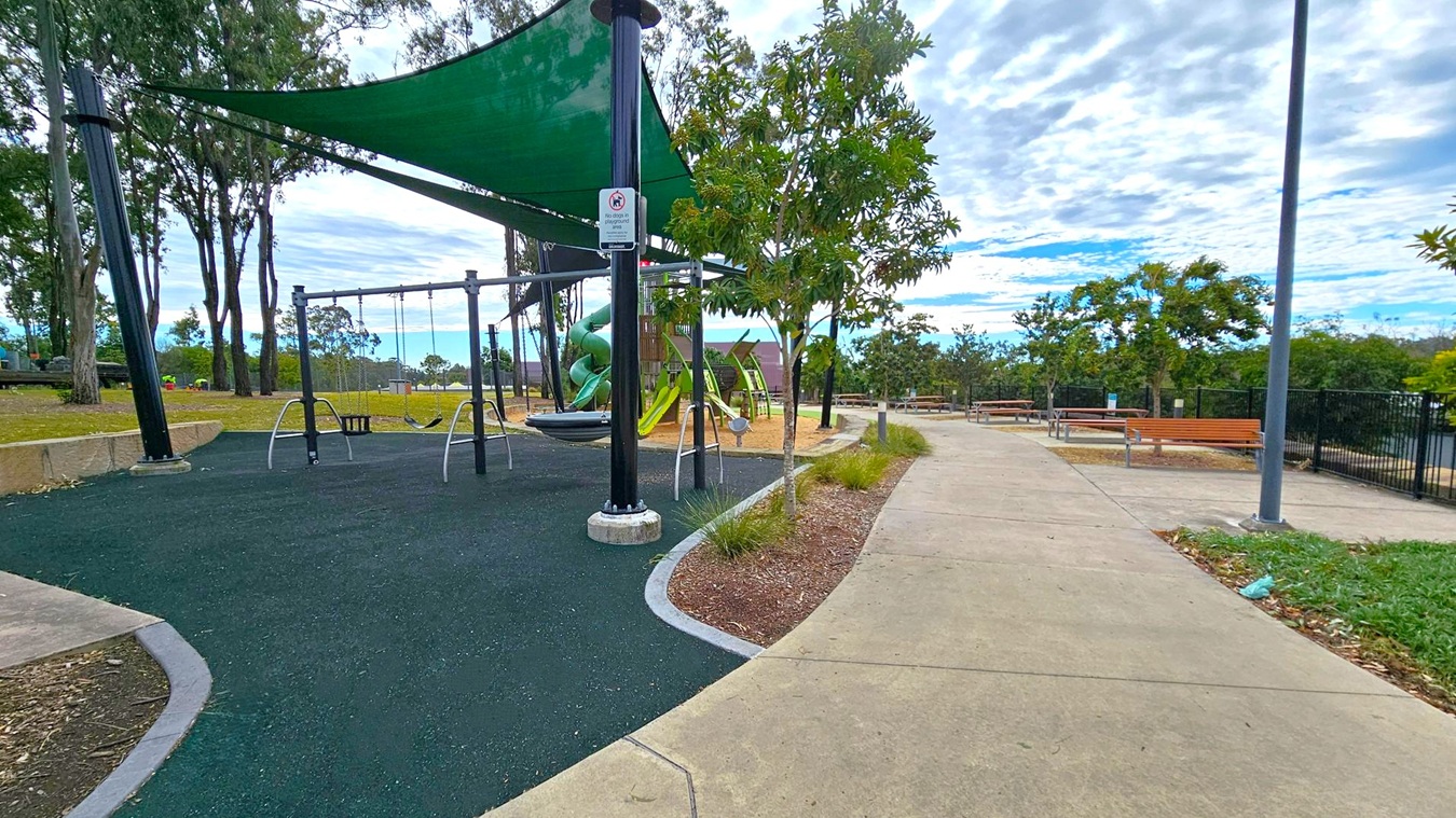 A playground with swings is on the left, covered by a green shade sail. The ground is covered with dark rubber matting. Behind the swings, there's a green spiral slide. Trees and a grassy area are visible in the background. On the right, a concrete path leads past wooden benches and a lamppost. The path is bordered by small plants and a fence is visible further back. The sky is overcast with scattered clouds. A playground with swings is on the left, covered by a green shade sail. The ground is covered with dark rubber matting. Behind the swings, there's a green spiral slide. Trees and a grassy area are visible in the background. On the right, a concrete path leads past wooden benches and a lamppost. The path is bordered by small plants and a fence is visible further back. The sky is overcast with scattered clouds.