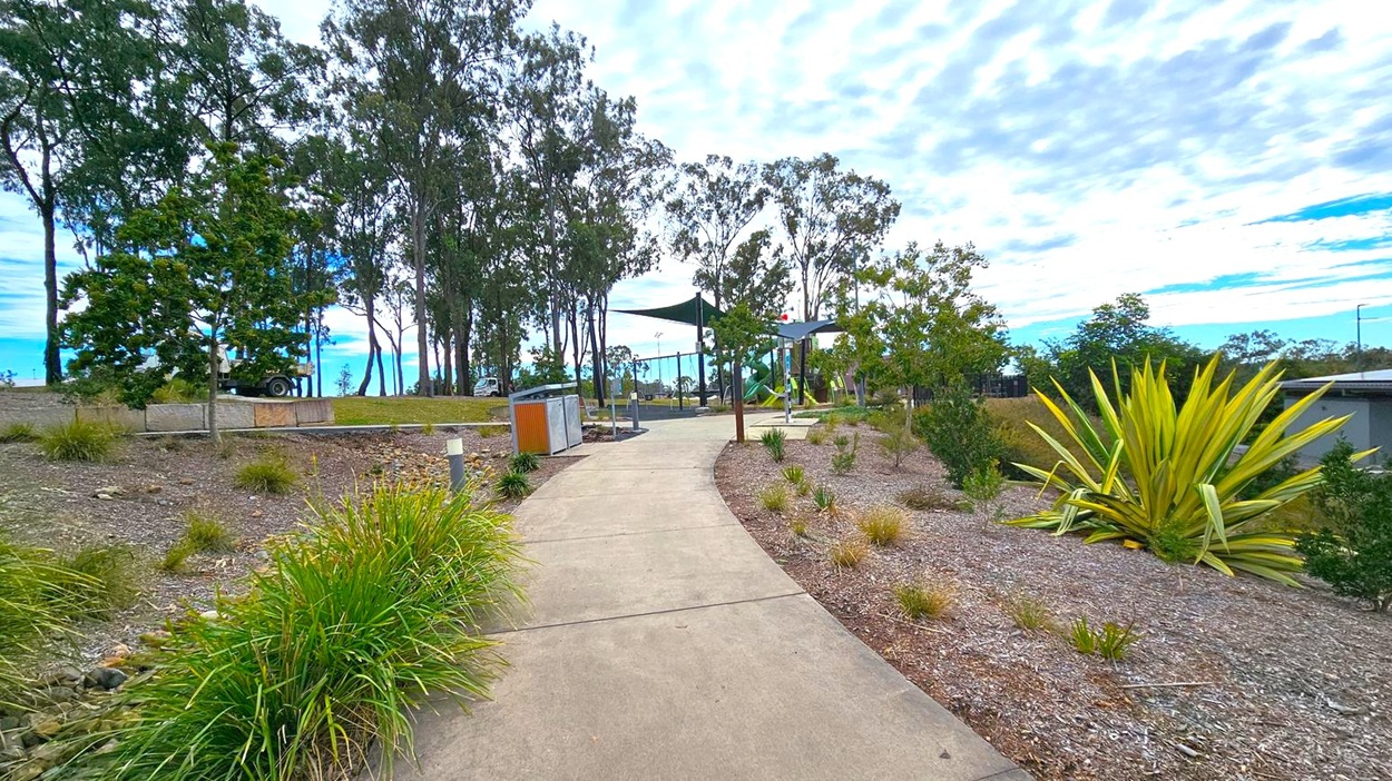 A winding concrete path leads through a landscaped area with various plants and shrubs on both sides. On the left, there are tall trees and a parked vehicle partially visible. To the right, a large spiky plant is prominent among smaller bushes. In the middle distance, a playground with green shade sails and equipment is visible. The ground is covered with mulch and gravel, and the sky is overcast with scattered clouds. A winding concrete path leads through a landscaped area with various plants and shrubs on both sides. On the left, there are tall trees and a parked vehicle partially visible. To the right, a large spiky plant is prominent among smaller bushes. In the middle distance, a playground with green shade sails and equipment is visible. The ground is covered with mulch and gravel, and the sky is overcast with scattered clouds.