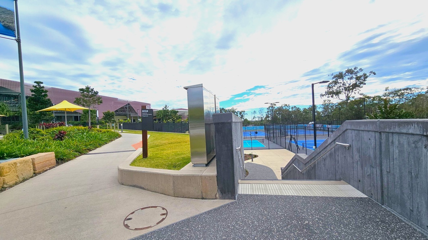 A pathway leads through a landscaped area with green grass and plants on the left. A yellow umbrella provides shade near a building with a sloped roof. On the right, a set of stairs descends to a series of blue tennis courts enclosed by fencing. Metal railings line the stairs, and tall trees are visible in the background. The sky is overcast with patches of blue. A signpost and a metallic structure are near the path. A pathway leads through a landscaped area with green grass and plants on the left. A yellow umbrella provides shade near a building with a sloped roof. On the right, a set of stairs descends to a series of blue tennis courts enclosed by fencing. Metal railings line the stairs, and tall trees are visible in the background. The sky is overcast with patches of blue. A signpost and a metallic structure are near the path.