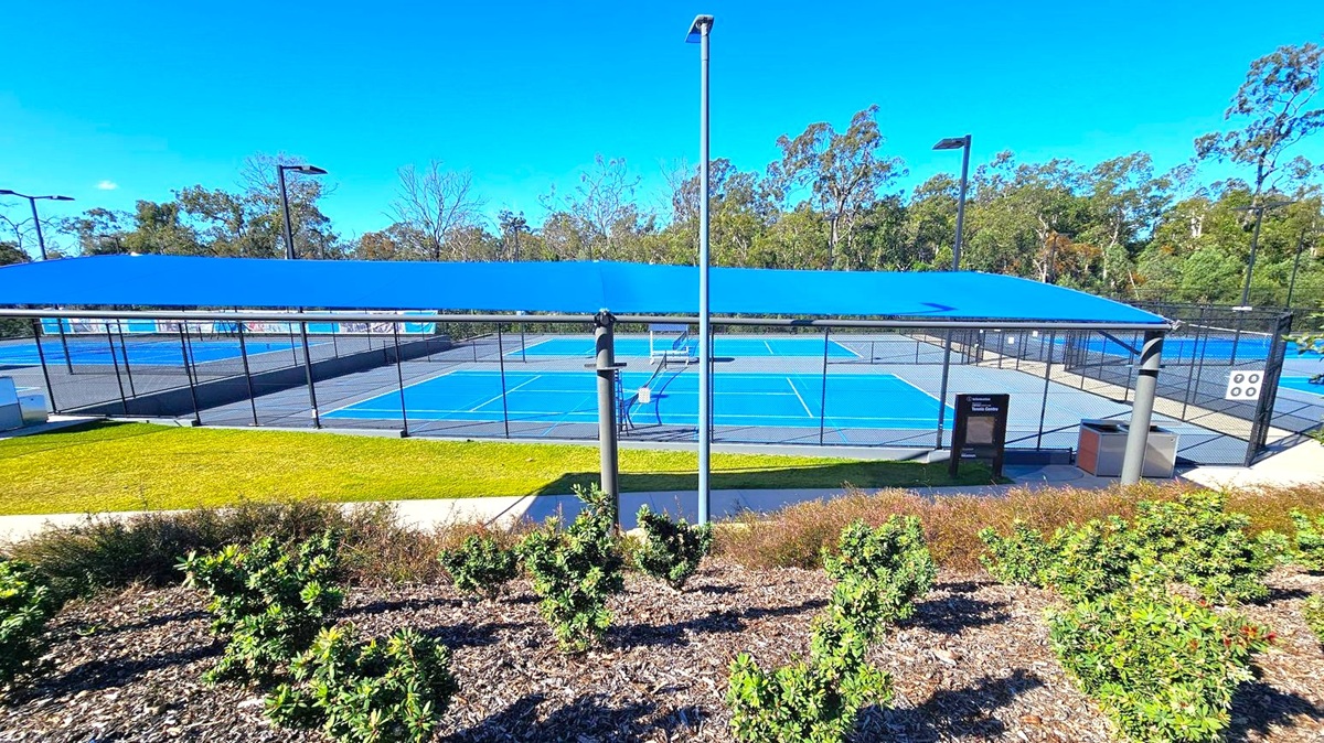 A tennis court complex with multiple blue courts is visible, surrounded by black chain-link fencing. A blue shade structure covers part of the area. In the foreground, there are small green shrubs and a patch of grass. Tall light poles are positioned around the courts. Trees and greenery form the background, with a clear blue sky above. A pathway runs parallel to the courts, and a sign and bin are near the entrance. A tennis court complex with multiple blue courts is visible, surrounded by black chain-link fencing. A blue shade structure covers part of the area. In the foreground, there are small green shrubs and a patch of grass. Tall light poles are positioned around the courts. Trees and greenery form the background, with a clear blue sky above. A pathway runs parallel to the courts, and a sign and bin are near the entrance.