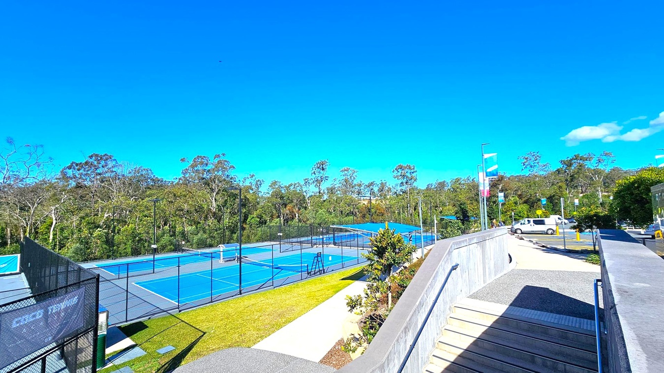 On the left, there are blue tennis courts surrounded by black fencing, with trees in the background. In the middle, a pathway with a small garden area runs alongside the courts. On the right, a concrete ramp with metal railings leads down to a car park where a white van is parked. Banners on poles line the path, and more trees are visible in the distance. The ground is a mix of grass, concrete, and asphalt. On the left, there are blue tennis courts surrounded by black fencing, with trees in the background. In the middle, a pathway with a small garden area runs alongside the courts. On the right, a concrete ramp with metal railings leads down to a car park where a white van is parked. Banners on poles line the path, and more trees are visible in the distance. The ground is a mix of grass, concrete, and asphalt.