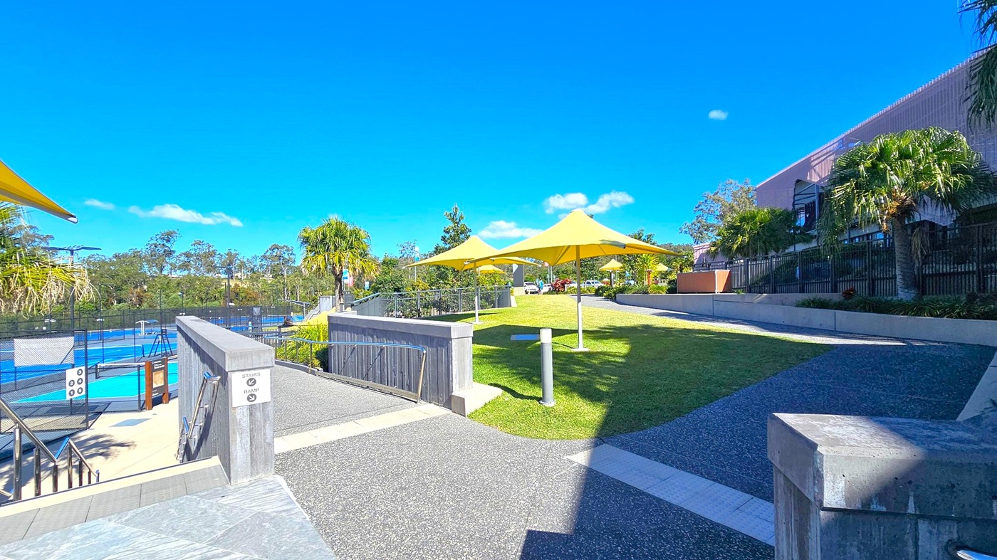A paved pathway leads through a landscaped area with green grass and several yellow shade structures. On the left, there is a fenced area with blue sports courts. The right side features a building with a modern design and palm trees along the perimeter. The background is filled with trees and a clear blue sky. A paved pathway leads through a landscaped area with green grass and several yellow shade structures. On the left, there is a fenced area with blue sports courts. The right side features a building with a modern design and palm trees along the perimeter. The background is filled with trees and a clear blue sky.