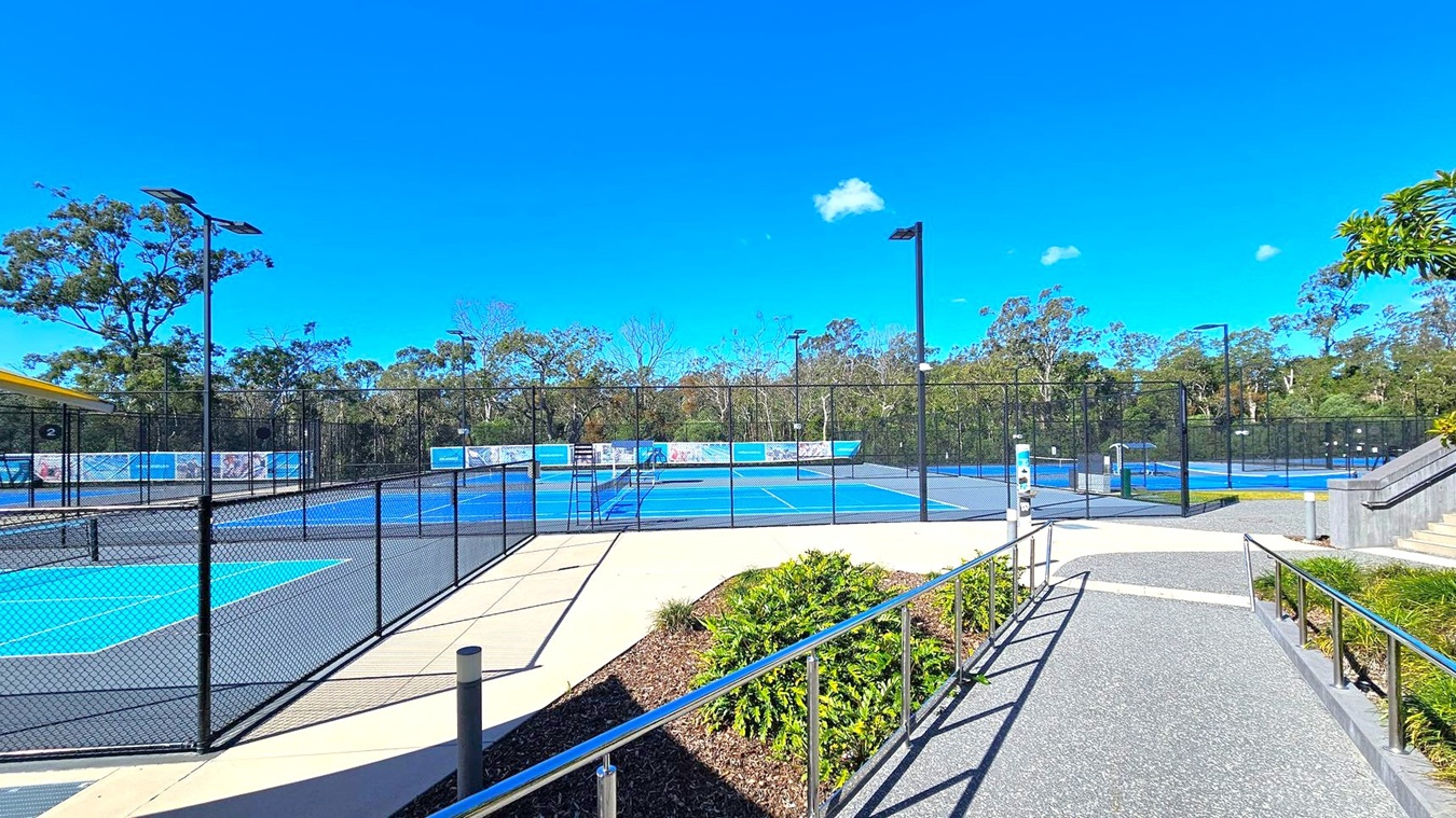 A tennis facility with multiple blue courts surrounded by black chain-link fencing. In the foreground, a pathway with metal railings leads towards the courts. On the right, a set of stairs is visible, bordered by greenery. The background features tall trees and a clear blue sky. A yellow-roofed structure is partially visible on the left. A tennis facility with multiple blue courts surrounded by black chain-link fencing. In the foreground, a pathway with metal railings leads towards the courts. On the right, a set of stairs is visible, bordered by greenery. The background features tall trees and a clear blue sky. A yellow-roofed structure is partially visible on the left.