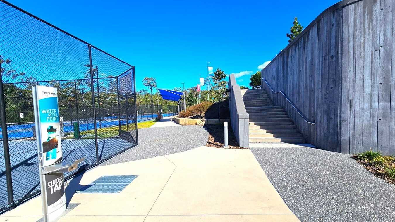 On the left, a chain-link fence encloses blue tennis courts. A water refill station with signage is nearby. In the centre, a paved path leads towards the courts and a grassy area. On the right, a tall concrete wall borders a set of stairs with a metal handrail, leading upwards. Trees and shrubs are visible in the background, and the ground is a mix of concrete and gravel. On the left, a chain-link fence encloses blue tennis courts. A water refill station with signage is nearby. In the centre, a paved path leads towards the courts and a grassy area. On the right, a tall concrete wall borders a set of stairs with a metal handrail, leading upwards. Trees and shrubs are visible in the background, and the ground is a mix of concrete and gravel.