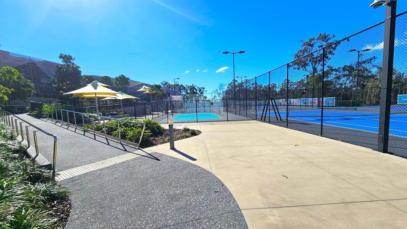 A pathway leads to a fenced tennis court area. On the left, there is a sloped walkway with metal railings and greenery alongside it. Yellow and white umbrellas provide shade over a seating area. On the right, the blue tennis courts are enclosed by a tall chain-link fence. Trees and a building are visible in the background. The ground is a mix of textured and smooth concrete. A pathway leads to a fenced tennis court area. On the left, there is a sloped walkway with metal railings and greenery alongside it. Yellow and white umbrellas provide shade over a seating area. On the right, the blue tennis courts are enclosed by a tall chain-link fence. Trees and a building are visible in the background. The ground is a mix of textured and smooth concrete.