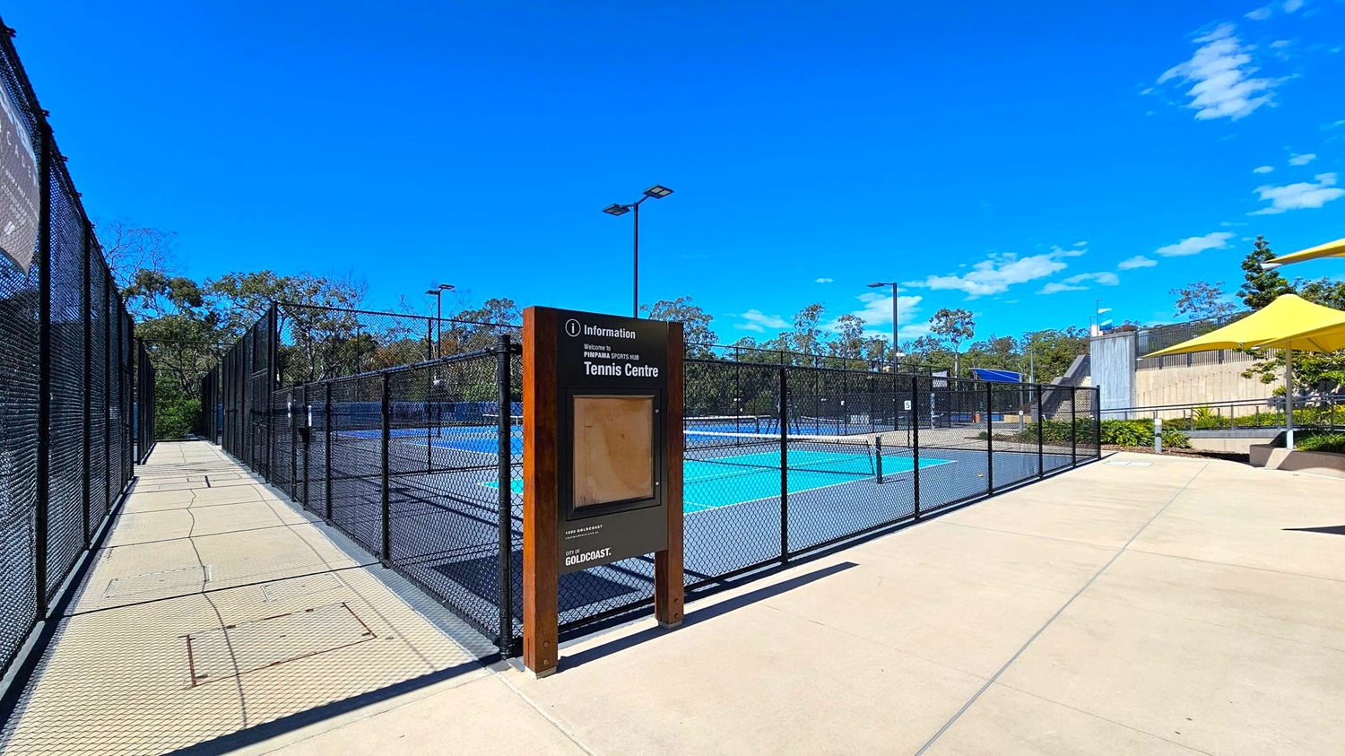 A tennis centre with a fenced court in the middle. On the left, a pathway runs alongside the fence, bordered by trees in the background. In the centre, a signboard provides information about the tennis centre. On the right, a concrete area with a yellow shade structure and some greenery. The ground is paved with light-coloured tiles, and tall lights are visible around the court. A tennis centre with a fenced court in the middle. On the left, a pathway runs alongside the fence, bordered by trees in the background. In the centre, a signboard provides information about the tennis centre. On the right, a concrete area with a yellow shade structure and some greenery. The ground is paved with light-coloured tiles, and tall lights are visible around the court.