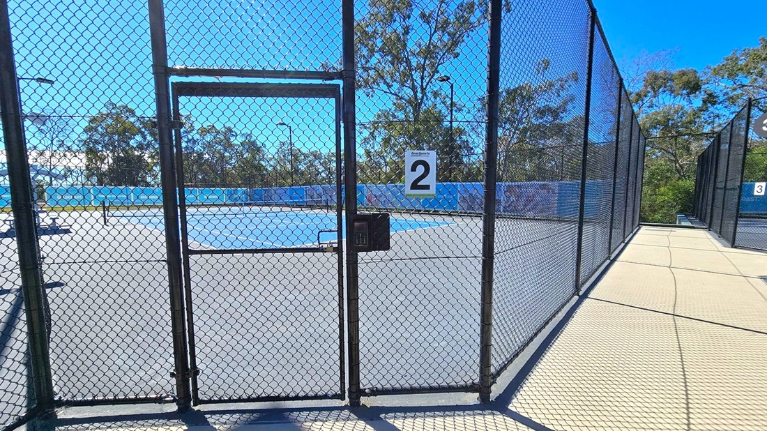 A tennis court enclosed by a chain-link fence with a gate in the centre, marked with the number "2". The court surface is blue and grey. To the right, a pathway runs alongside the fence, leading to another court marked "3". In the background, there are trees and a blue sky. The ground outside the court is a light-coloured concrete. A tennis court enclosed by a chain-link fence with a gate in the centre, marked with the number "2". The court surface is blue and grey. To the right, a pathway runs alongside the fence, leading to another court marked "3". In the background, there are trees and a blue sky. The ground outside the court is a light-coloured concrete.