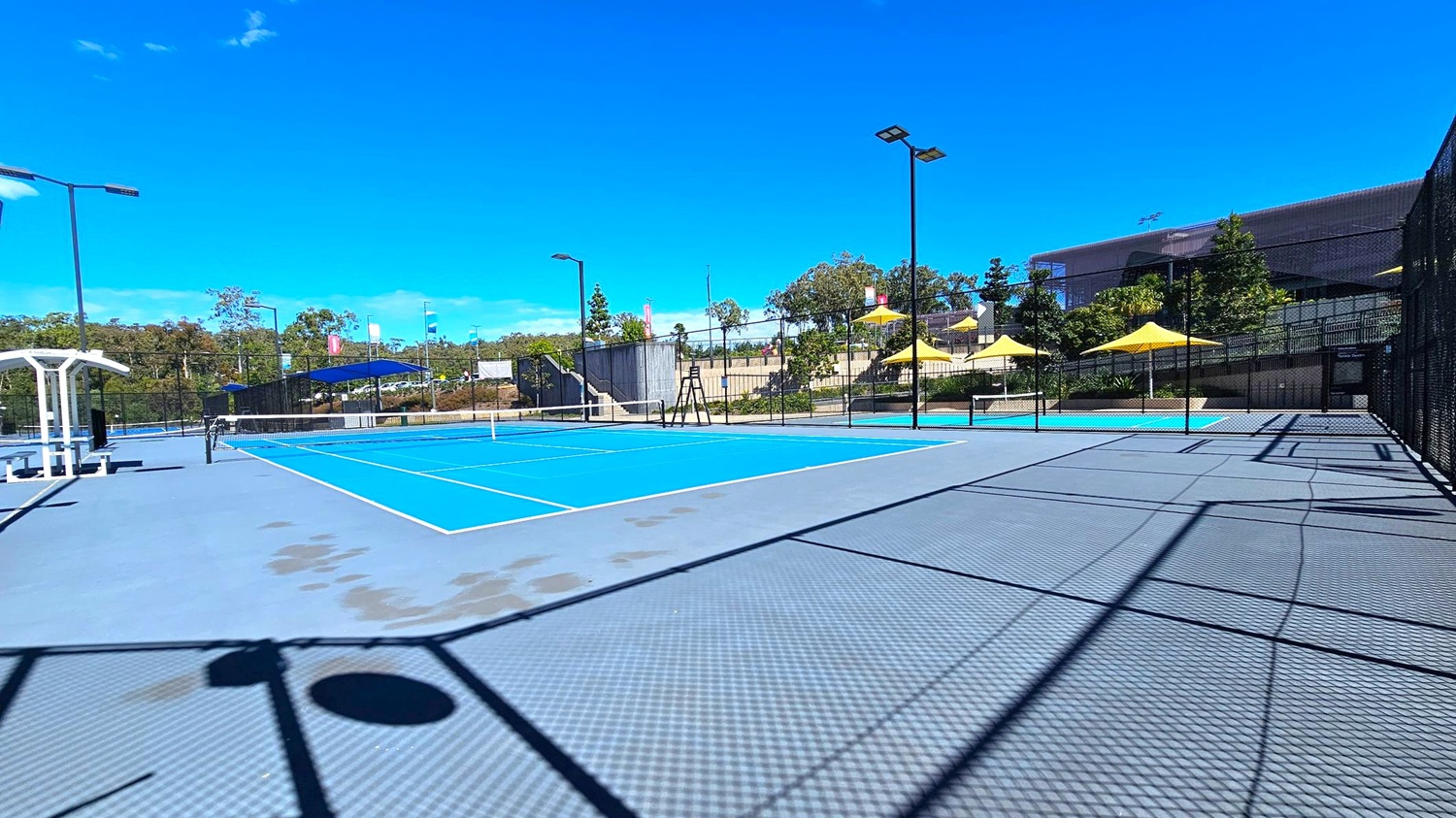 A tennis court with a bright blue playing surface and grey surrounding area. On the left, there is a white bench with a canopy. The court is enclosed by a black chain-link fence. In the background, there are several yellow umbrellas providing shade, and a building partially obscured by trees. Tall floodlights are positioned around the court. The ground is patterned with shadows from the fence and other structures. A tennis court with a bright blue playing surface and grey surrounding area. On the left, there is a white bench with a canopy. The court is enclosed by a black chain-link fence. In the background, there are several yellow umbrellas providing shade, and a building partially obscured by trees. Tall floodlights are positioned around the court. The ground is patterned with shadows from the fence and other structures.