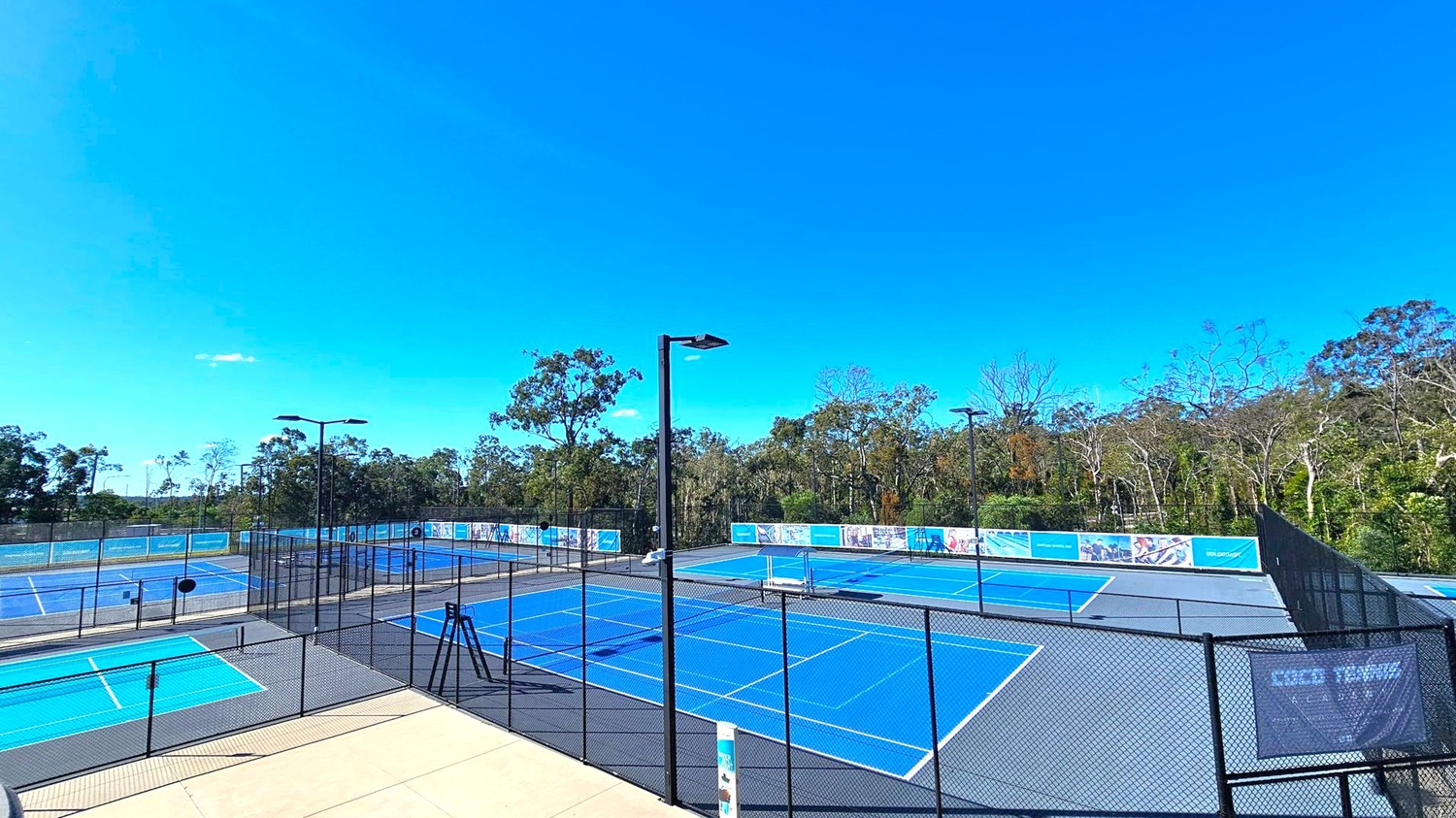 Tennis courts with blue surfaces are surrounded by black chain-link fencing. Several tall floodlights are positioned around the courts. In the background, there is a dense line of trees and vegetation. The courts are empty, and there are banners on the fences. The ground is a mix of concrete and court surfaces. Tennis courts with blue surfaces are surrounded by black chain-link fencing. Several tall floodlights are positioned around the courts. In the background, there is a dense line of trees and vegetation. The courts are empty, and there are banners on the fences. The ground is a mix of concrete and court surfaces.