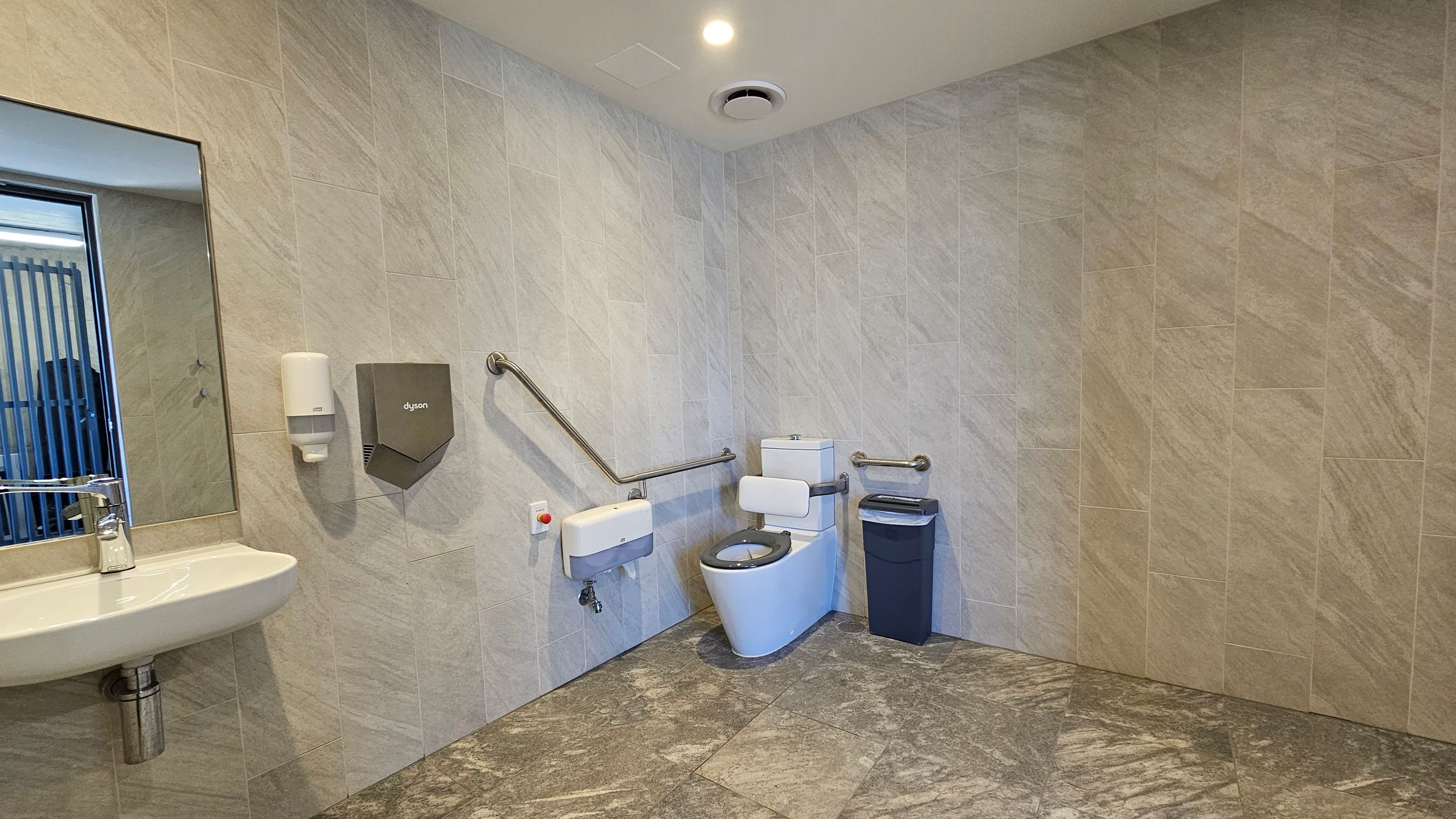A spacious, accessible bathroom with light-coloured tiled walls and a patterned floor. On the left, a wall-mounted sink with a mirror above it, accompanied by a soap dispenser and a hand dryer. In the centre, a toilet with support rails on both sides and a small bin to the right. The ceiling features a light and a ventilation fan. A spacious, accessible bathroom with light-coloured tiled walls and a patterned floor. On the left, a wall-mounted sink with a mirror above it, accompanied by a soap dispenser and a hand dryer. In the centre, a toilet with support rails on both sides and a small bin to the right. The ceiling features a light and a ventilation fan.