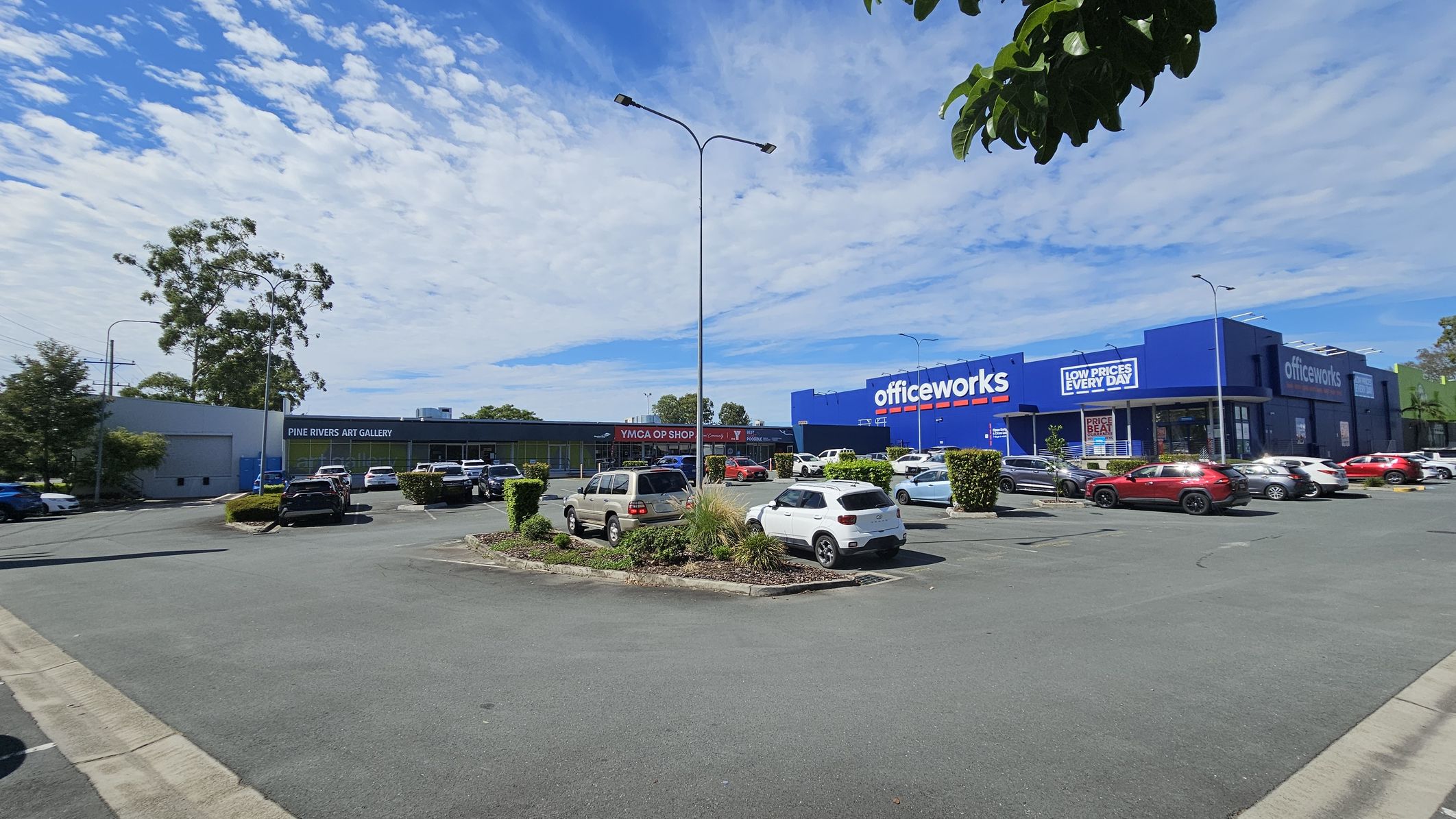 A car park with several vehicles is in the foreground, surrounded by small bushes and trees. On the left, a building with a sign for "Pine Rivers Art Gallery" is visible, alongside a "YMCA Op Shop". On the right, a large blue building with "Officeworks" signage is prominent. The background features tall trees and a partly cloudy sky. A few streetlights are scattered throughout the area.