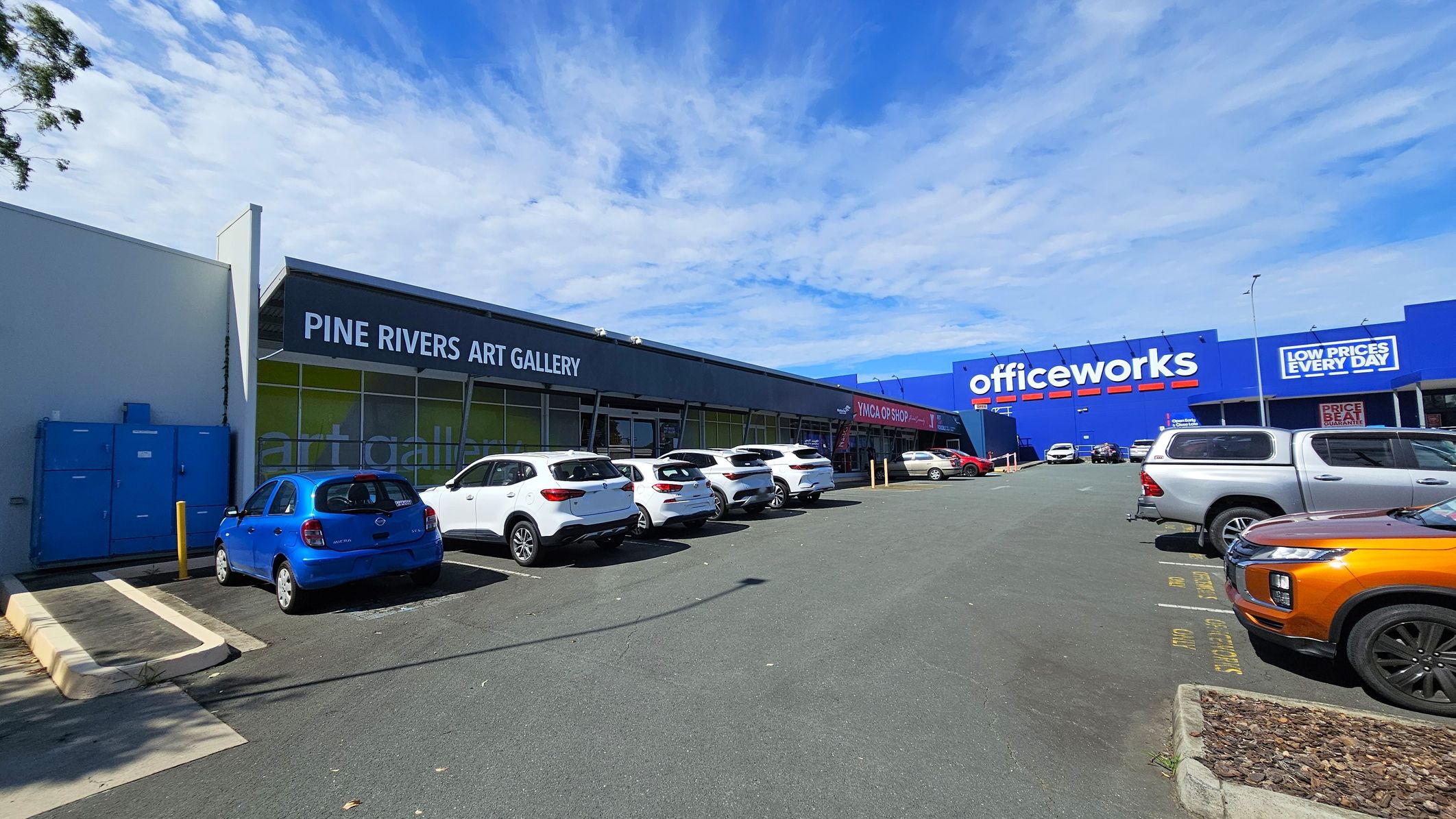 On the left, a row of parked cars is in front of the Pine Rivers Art Gallery, with a blue car closest to the camera. The building has a dark facade with large windows. On the right, a large blue Officeworks building is visible, with a sign advertising "Low Prices Every Day." Several cars are parked in front, including a silver utility vehicle and an orange car. The ground is asphalt, and there are some small patches of landscaping.
