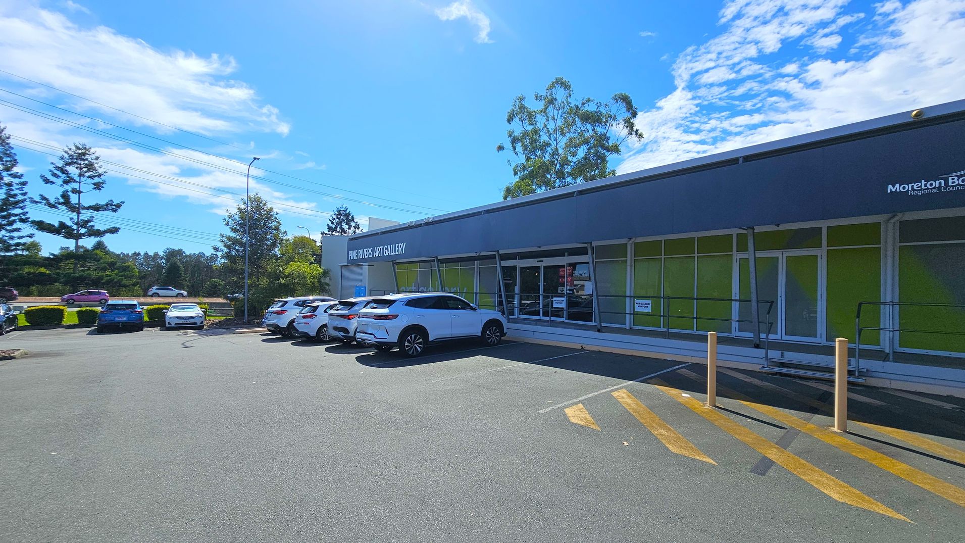 A modern building with a grey facade and large windows on the right, labelled "Pine Rivers Art Gallery." Several white cars are parked in front of the building. The foreground features a paved area with yellow diagonal lines and bollards. In the background, there are trees and a road with a few vehicles. The sky is clear with some clouds.
