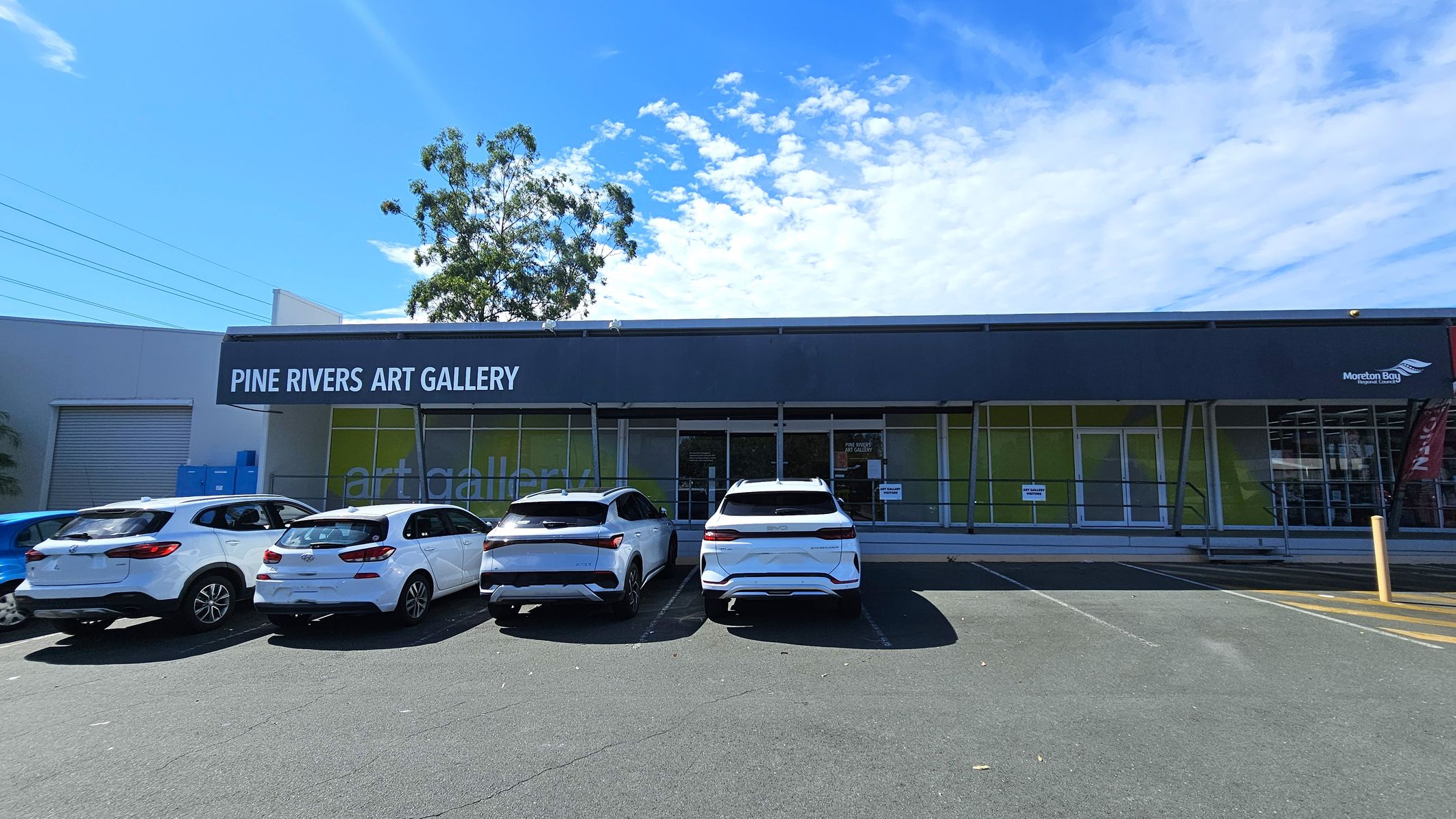 A row of four white cars is parked in front of a building with a dark blue awning that reads "Pine Rivers Art Gallery." The building has large glass windows with green panels behind them. A tree is visible to the left, and there are some blue utility boxes near the building. The ground is an asphalt parking lot with marked spaces.