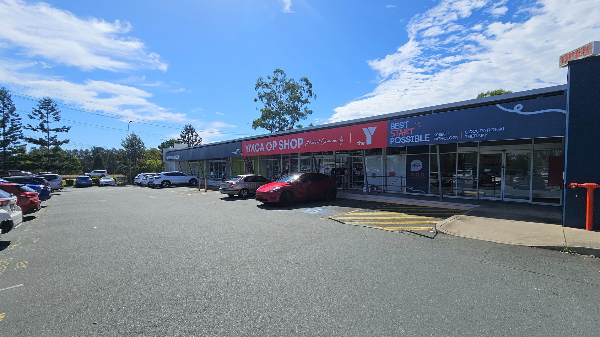 A car park with several vehicles on the left, including a red car near the entrance of a building. The building on the right has signage for "YMCA OP SHOP" and "BEST START POSSIBLE" with services like speech pathology and occupational therapy. The foreground shows a paved area with a yellow marked zone. Trees are visible in the background, and the sky is clear with some clouds.