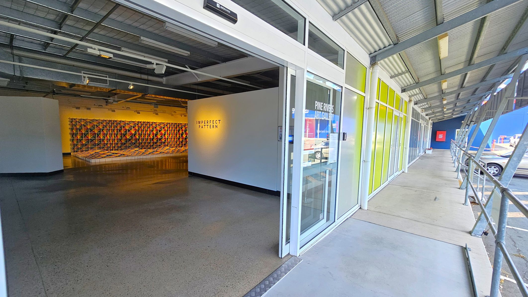 Entrance to an art gallery with a glass door on the right, labelled "Pine Rivers Art Gallery". The exterior features a covered walkway with a metal roof and railing. Inside, a wall displays the text "IMPERFECT PATTERN" with a geometric art installation in the background. The floor is polished concrete, and the ceiling has exposed beams and lighting fixtures.