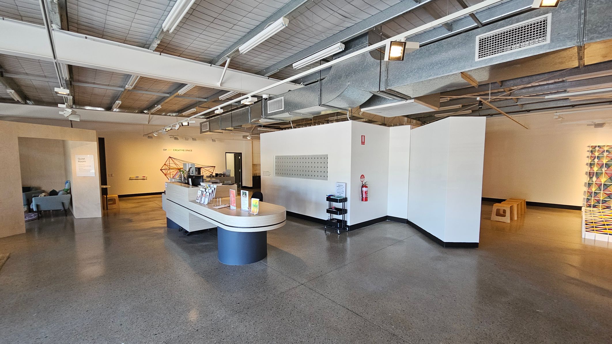 A modern interior space with a polished concrete floor and an industrial-style ceiling featuring exposed ducts and lighting. In the centre, there's a curved reception desk with brochures and pamphlets. To the left, a quiet room with a sofa is visible. On the right, a wall with a fire extinguisher and a small black trolley. In the background, a creative space with geometric wall art and wooden stools.