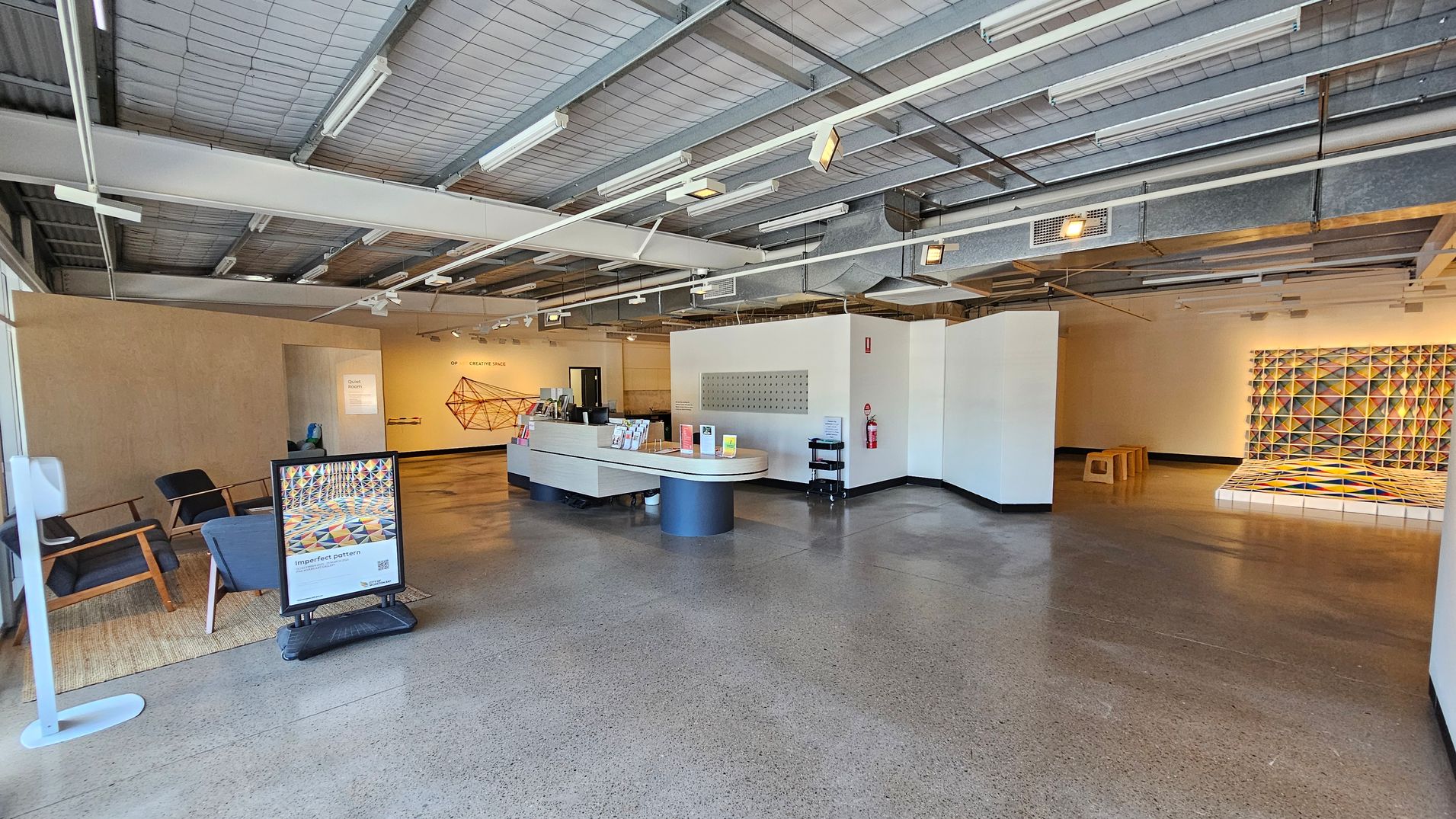 A spacious interior with a polished concrete floor and an industrial-style ceiling with exposed beams and lighting fixtures. On the left, there are two chairs on a rug and a stand with a colourful poster. In the centre, a curved reception desk displays brochures. The right side features a geometric art installation on the wall, with a small wooden bench nearby. Walls are mostly white, and a fire extinguisher is mounted on one.