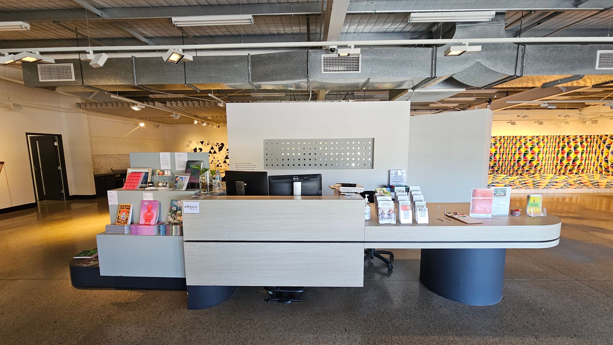 A modern reception area with a light-coloured desk in the centre, displaying brochures, pamphlets, and a card machine. Behind the desk, there are two computer monitors. The floor is polished concrete, and the ceiling features exposed ductwork and lighting fixtures. On the left, a dark door is visible, while the right side showcases a vibrant, geometric wall mural with a pattern of colourful triangles.