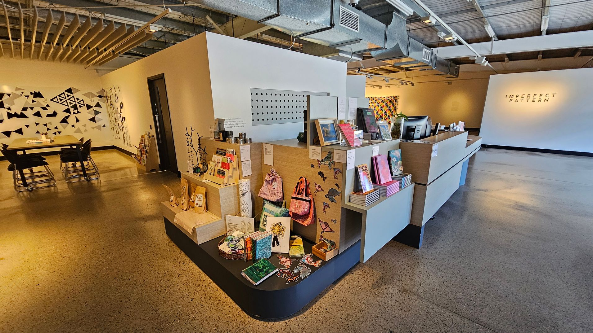 A modern interior with a polished concrete floor and an exposed ceiling with ductwork. On the right, a reception desk displays books and art items. In the centre, a wall features geometric art and a sign reading "IMPERFECT PATTERN." On the left, a table with chairs is set against a wall adorned with black and white triangular patterns. The space is well-lit with track lighting, creating a contemporary and artistic atmosphere.