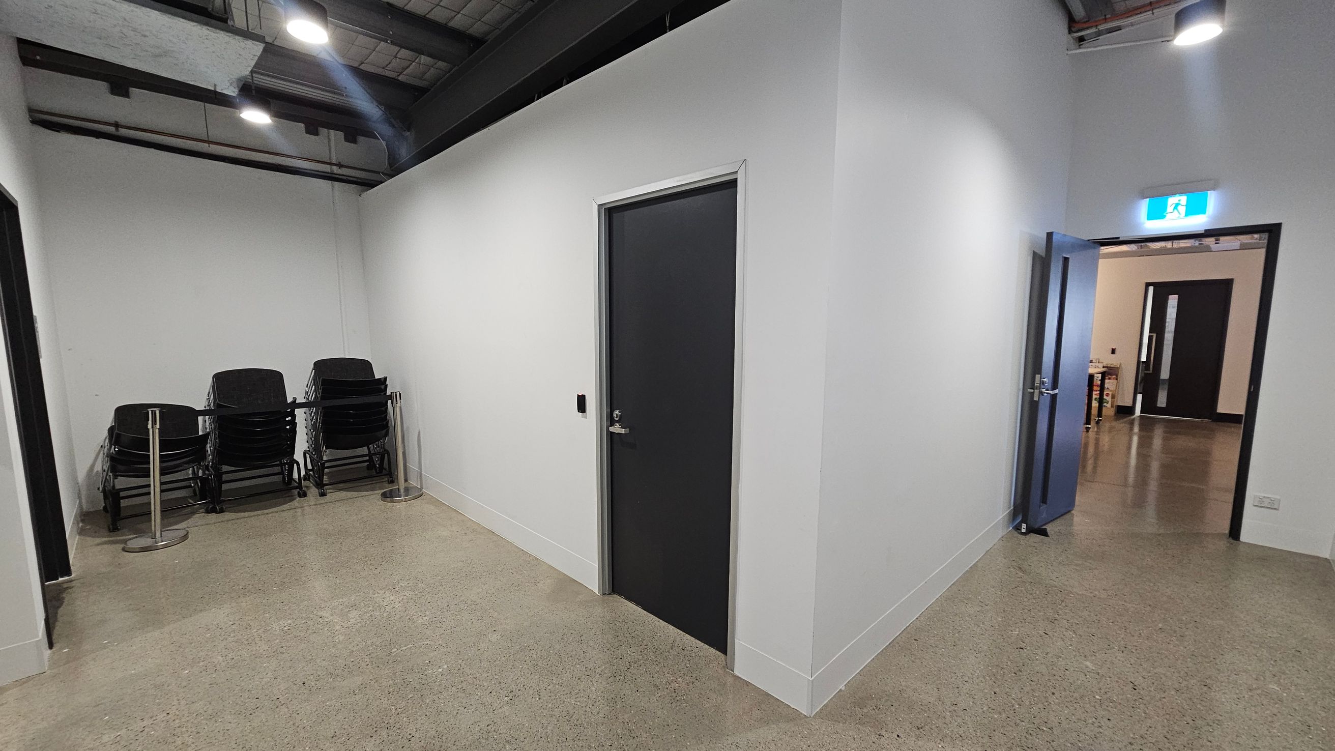 A hallway with polished concrete floors and white walls. On the left, there is a stack of black chairs against the wall, secured by a barrier. In the centre, a closed dark door is visible. On the right, an open door leads to another room, with an illuminated exit sign above it. The ceiling is industrial, featuring exposed beams and lighting fixtures.