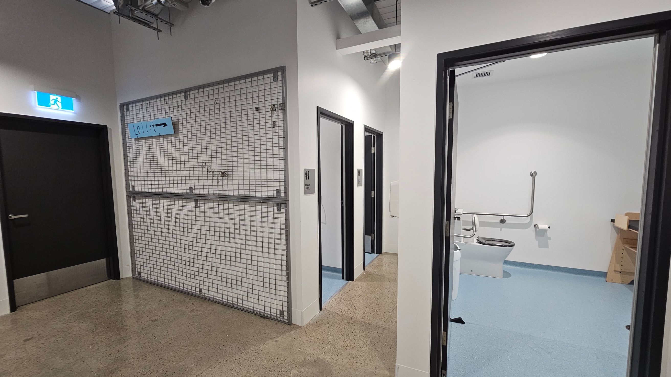 On the left, a black door with an illuminated exit sign above it. Next to it, a metal grid wall with a blue "toilet" sign. In the centre, two open doors with signs indicating male and female toilets. On the right, an open door reveals a bathroom with a toilet, grab bars, and a toilet roll holder. The floor is a mix of polished concrete and blue vinyl, and the ceiling has exposed ductwork.