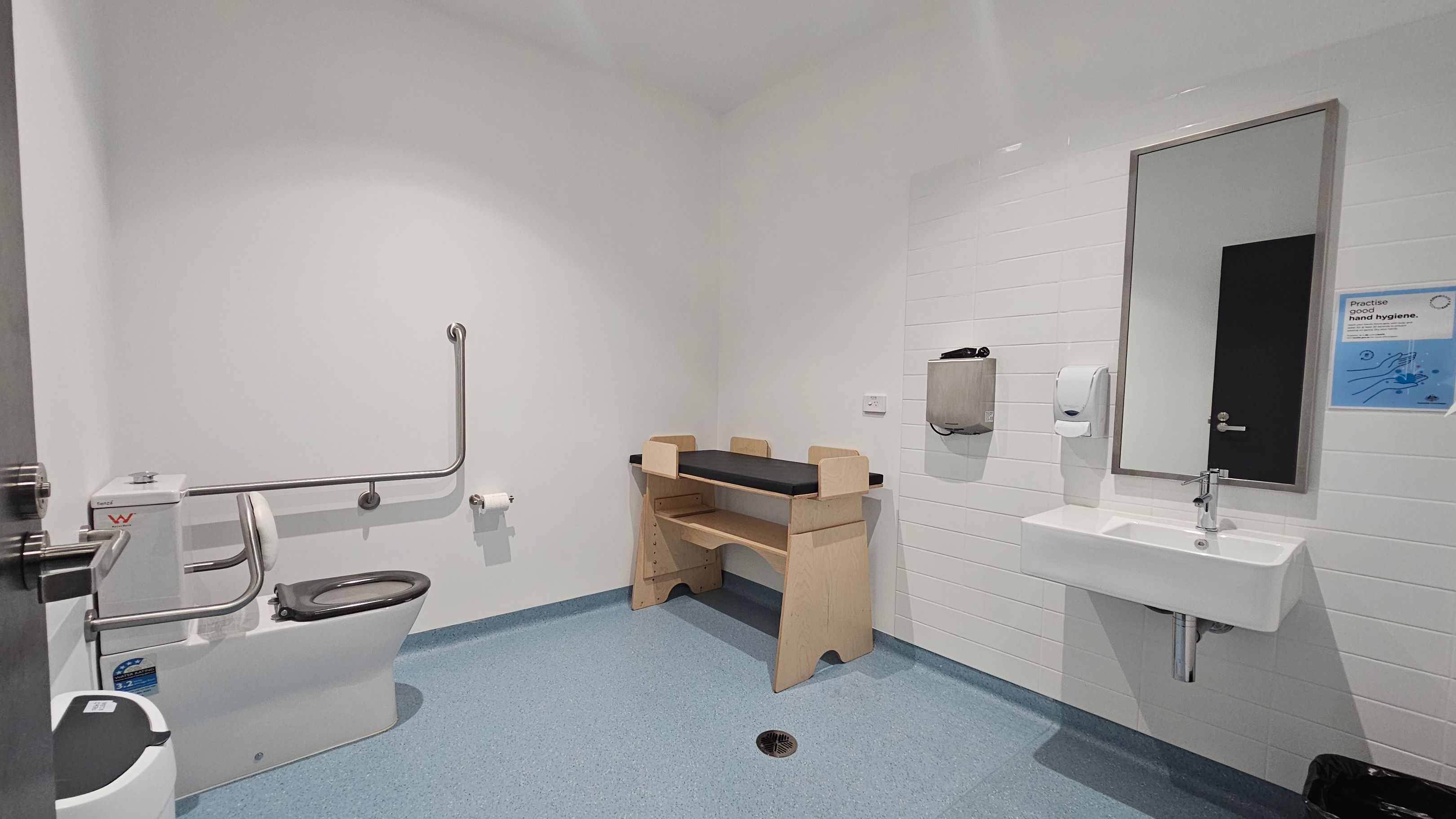 A public restroom with a blue floor and white walls. On the left, there is a toilet with a black seat, equipped with metal support bars and a toilet roll holder. In the centre, a wooden baby changing table with a black mat is positioned against the wall. On the right, a white sink with a mirror above it is mounted on the wall, accompanied by a soap dispenser and a hand dryer. A sign about hand hygiene is visible next to the mirror.