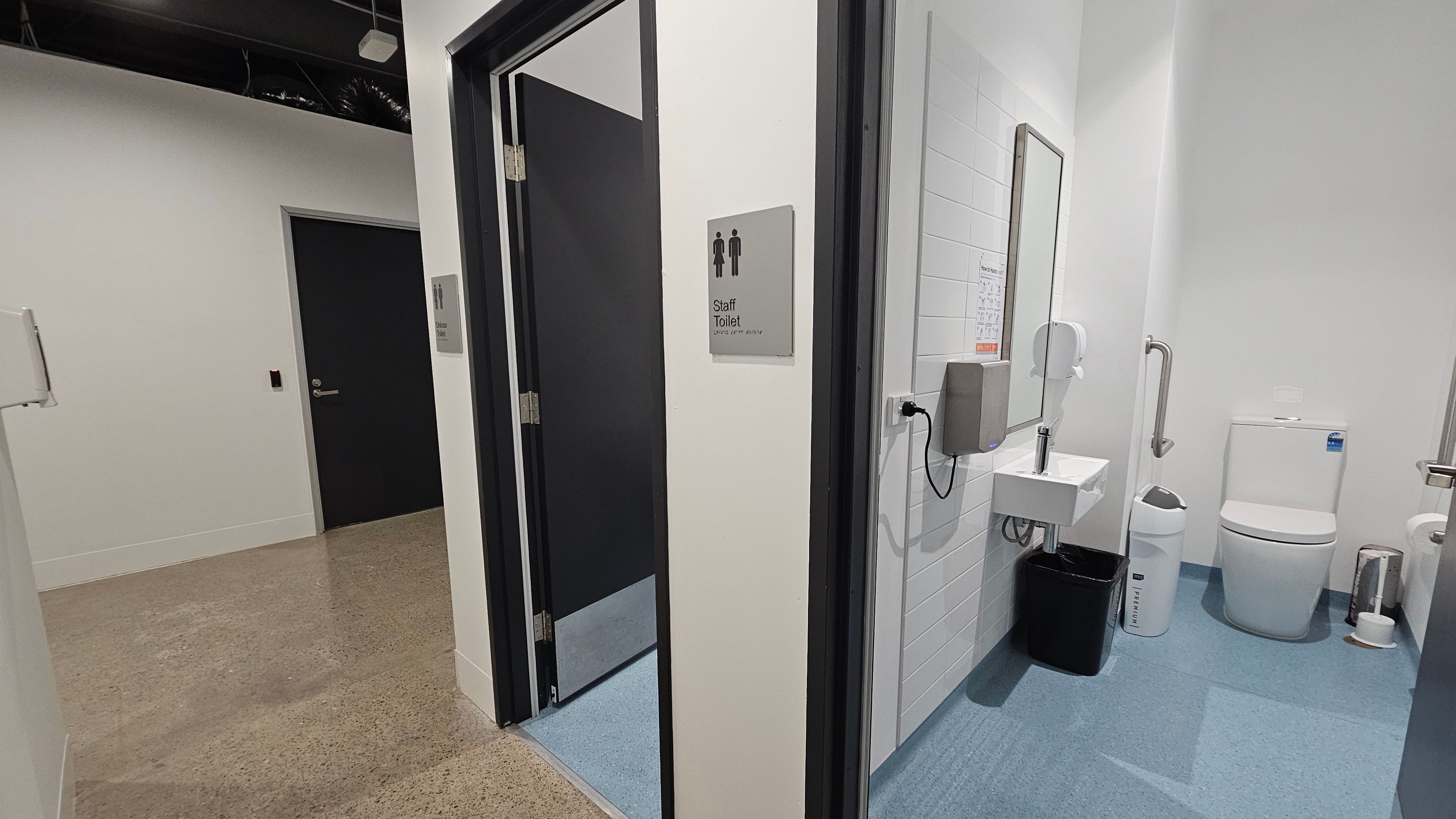 A corridor with a polished concrete floor leads to a staff toilet. The door to the toilet is open, revealing a room with a blue floor. Inside, there's a white toilet, a small sink with a mirror above it, and a wall-mounted soap dispenser. A black bin and a white sanitary bin are next to the sink. The walls are white with a tiled section near the sink. A black door is visible in the corridor, and the ceiling has exposed ductwork.
