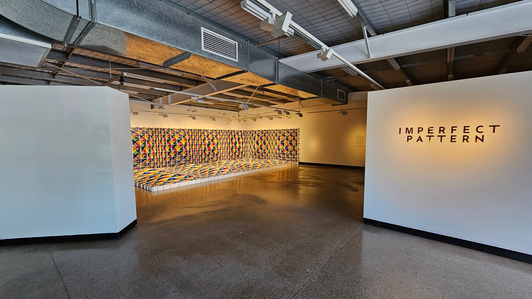 A gallery space with a polished concrete floor and an industrial-style ceiling with exposed ducts. On the right, a white wall displays the text "IMPERFECT PATTERN" in bold black letters. In the background, a vibrant, geometric mural covers the wall and floor, featuring a colourful pattern of triangles in various shades. The lighting is bright, highlighting the artwork and creating reflections on the floor.